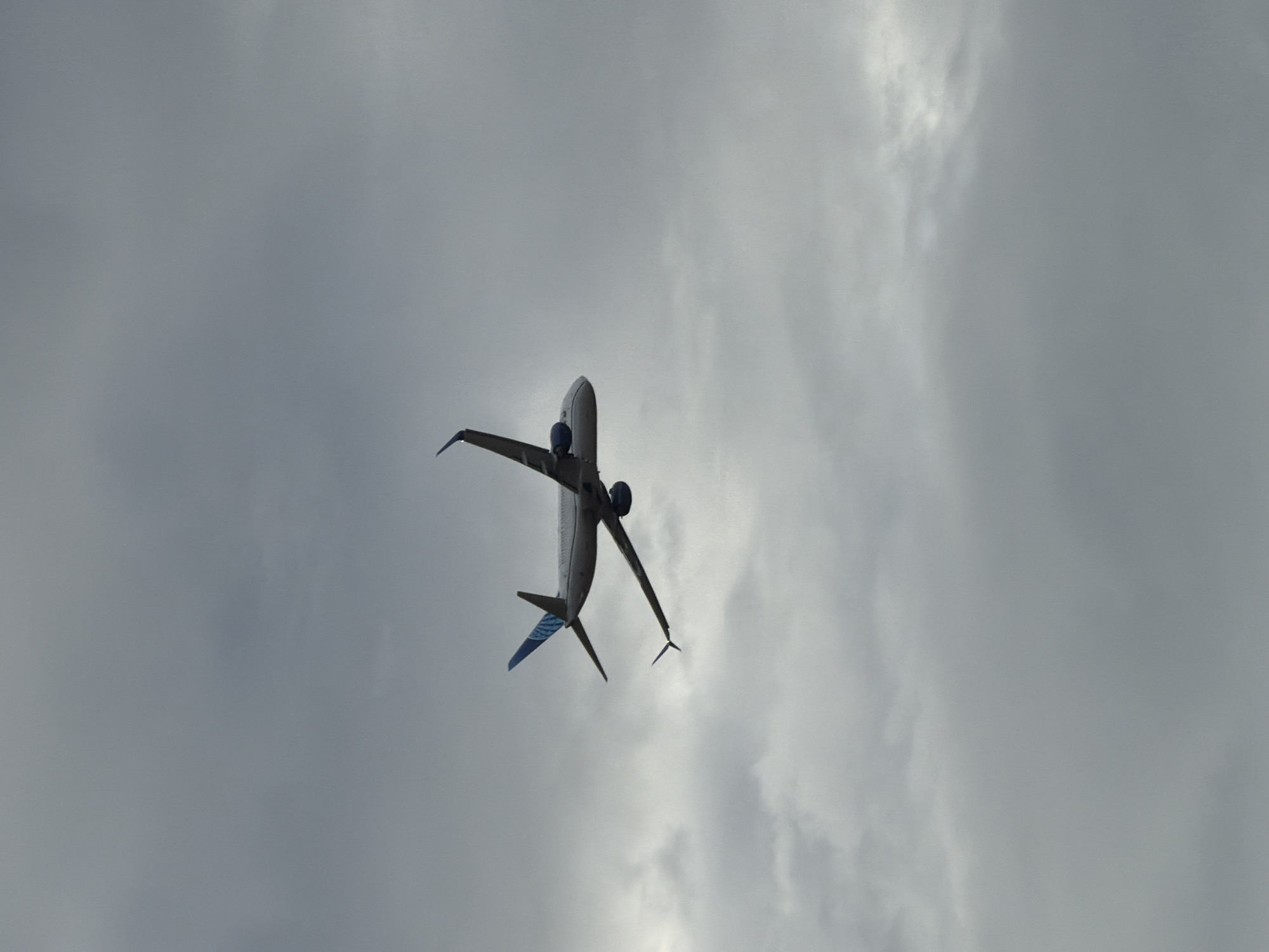 The image shows the underside of an airplane flying through a cloudy sky. The airplane is angled upwards, with its wings spread wide. The sky is overcast with thick gray clouds, suggesting it is a cloudy day. The lighting is diffuse, indicating it could be daytime, but the exact time is not discernible. The perspective is from below the aircraft, looking up. The airplane appears to be a commercial jetliner, with visible engines mounted under the wings and a tail with blue accents. There is no discernible text in the image. The scene is set against a backdrop of clouds, with no clear geographical features to indicate a specific location, though the context provided suggests Sacramento, United States. The overall mood is somewhat somber due to the overcast weather.