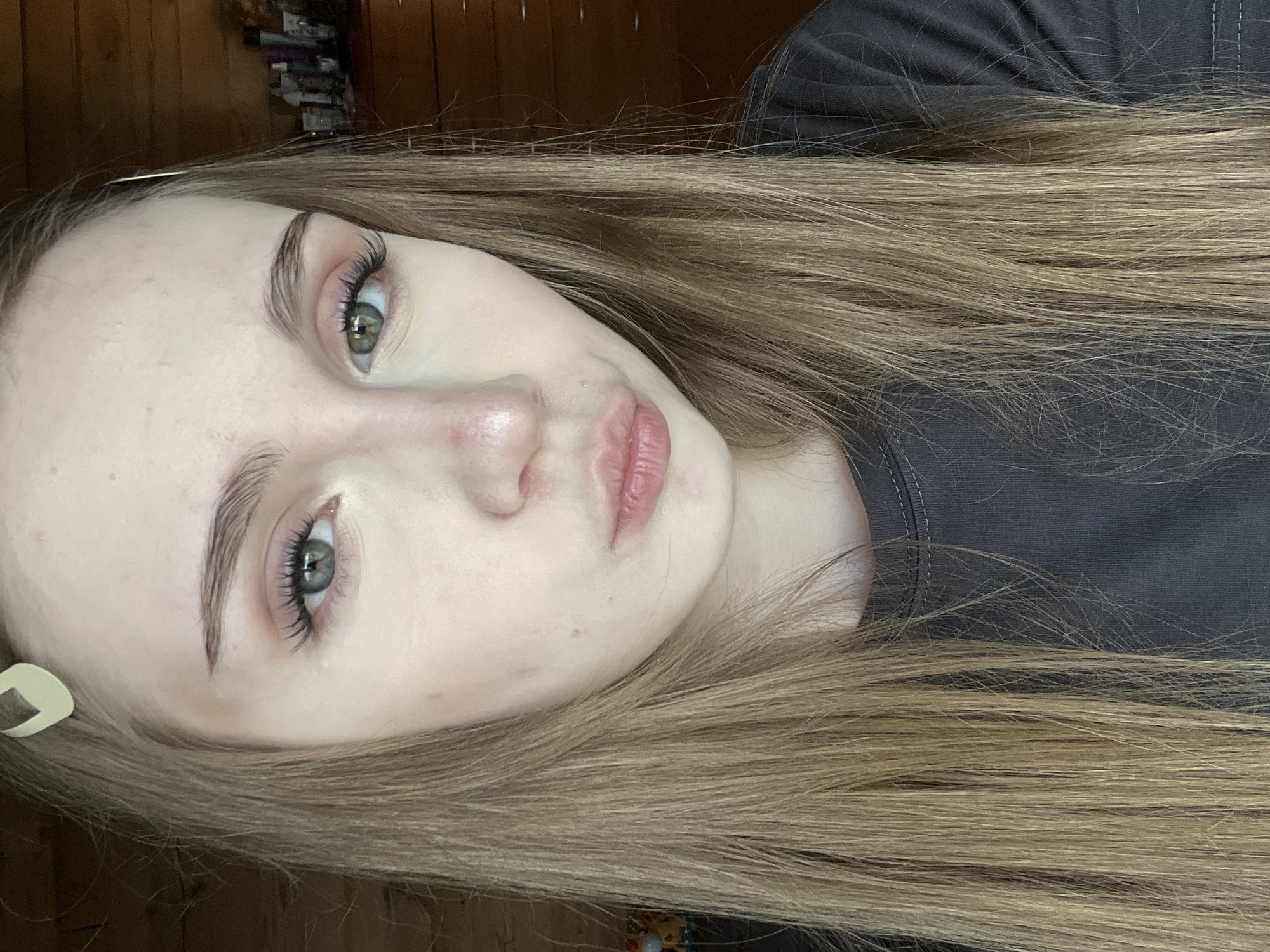 A young woman with long blonde hair and green eyes is looking at the camera. She is wearing a dark gray t-shirt and has a light-colored hair clip in her hair. Her makeup is subtle, with mascara and a light pink lipstick. The background appears to be an indoor setting with wooden walls and a dresser with various items on it. There is no discernible location in Ashkadar, Russia.