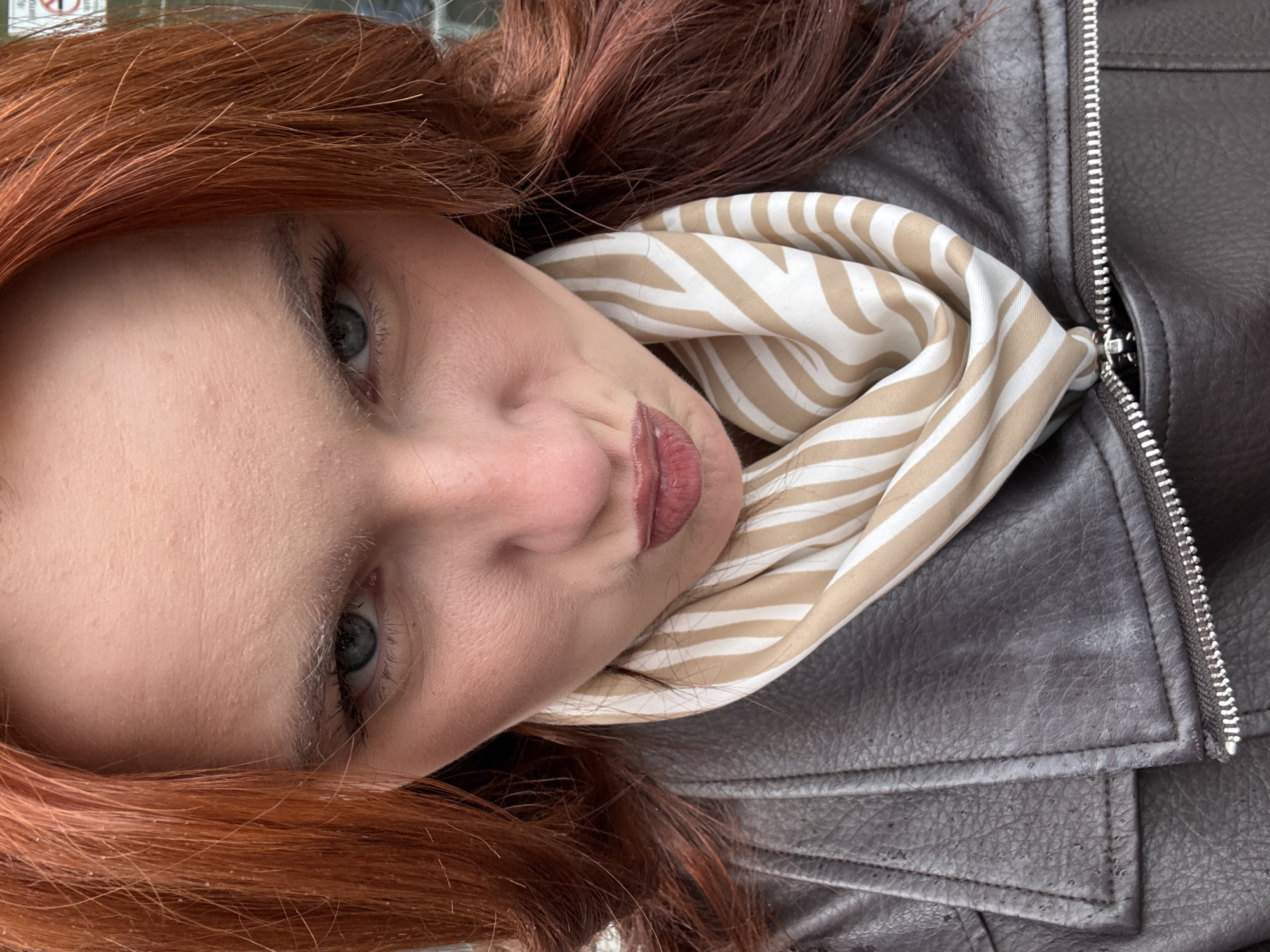 A close-up portrait depicts a fair-skinned woman with auburn hair and gray-blue eyes. Her face is slightly tilted, with her lips pursed in a subtle manner. She is wearing a brown leather jacket, the zipper visible on the right side of the frame. Around her neck, a beige and white striped scarf is draped. The image orientation is vertical, but the woman's face is angled towards the left. The environment is not specifically discernable, lacking elements clearly related to Saint Petersburg, Russia.
