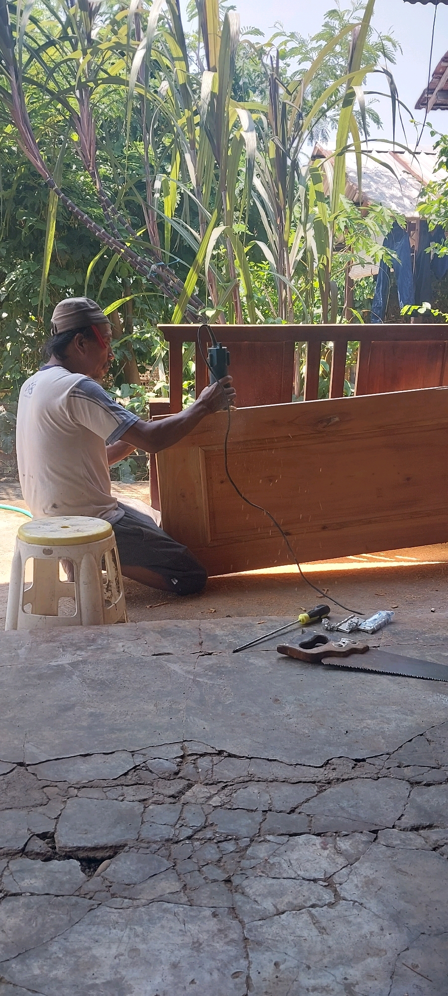 In Keling, Indonesia, a man is working on a wooden furniture piece outdoors. He is kneeling on the ground, using a green power tool, likely a router, on a large, unfinished wooden surface that appears to be part of a bench or bed frame. He is wearing a light-colored t-shirt, dark pants, and a beige head covering. To his left, a small white stool with a yellow seat is visible. The ground is a rough concrete surface with visible cracks. Tools, including a handsaw and a screwdriver, are lying on the ground near the furniture. In the background, tall green plants, possibly sugarcane, are growing, along with some distant structures and a blue tarp. The scene is illuminated by bright sunlight, casting shadows.