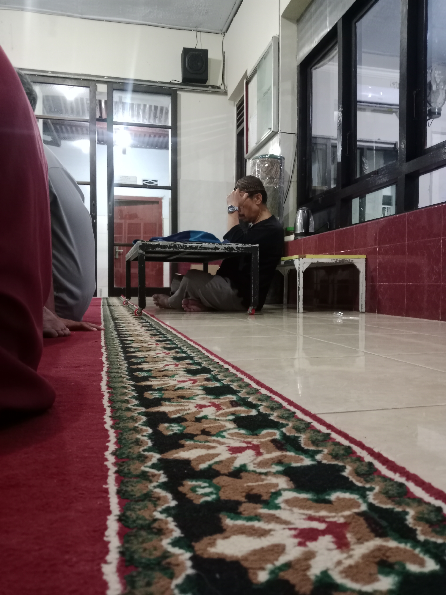 The image depicts an indoor scene, likely a place of worship or a communal gathering space, given the presence of prayer mats and a man in contemplative posture. The flooring is tiled, with a prominent red carpet featuring a patterned border in green, white, and brown. A black metal table with red wheels is positioned centrally. A man, wearing a peci (a type of cap) and dark clothing, sits on the floor, leaning forward with his hand on his forehead, appearing to be in deep thought or prayer. His right leg is crossed over his left. To the left and foreground, partially visible, are two other individuals, one in a dark maroon garment and another in a grey garment, also seated on the floor. The background reveals large glass sliding doors, offering a glimpse of what appears to be an outdoor or separate area with wooden doors. A black speaker is mounted high on the wall. The overall lighting suggests an indoor environment with artificial light. The scene is devoid of any specific contextual markers that would pinpoint its exact location within Kediri City, Indonesia, other than the assumption that this is an interior space within that city.
