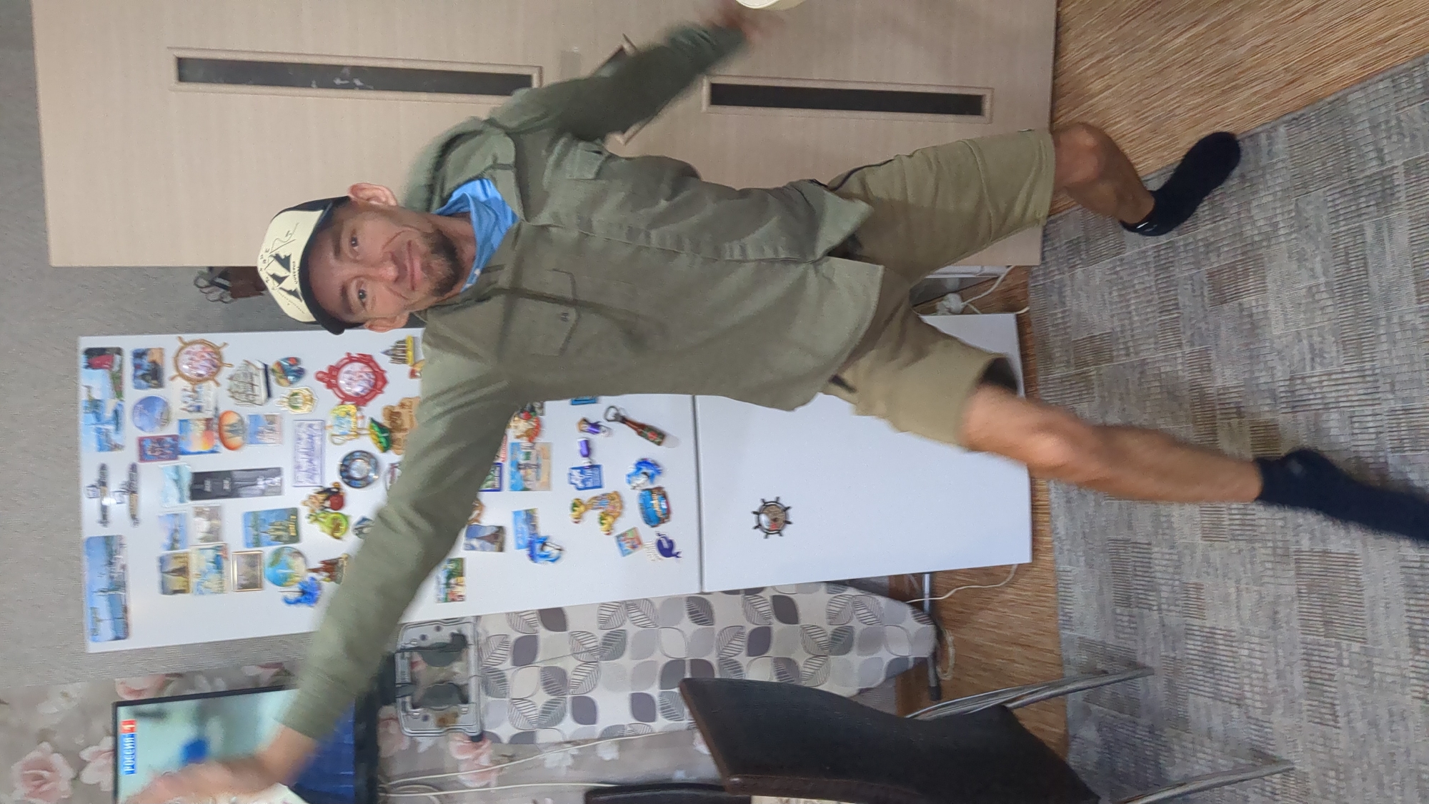 A full-body shot depicts a man indoors, likely in his home, exhibiting a playful pose. The man is wearing a light green button-up jacket over a blue shirt, khaki shorts, and a black and white baseball cap. He is wearing black socks on both feet.
Behind him is a white refrigerator covered with souvenir magnets. To the left of the refrigerator, a television is visible, displaying what appears to be Russian news. The background includes a door with horizontal rectangular details, a printed beige and white wallpaper, wooden flooring, and a gray rug. There are also chairs visible near the bottom of the image.
While not explicitly stated, the presence of Russian text on the television suggests a location within Russia. Without additional evidence, the specific city of Ulyanovsk cannot be confirmed. The image depicts a casual scene within a domestic environment, focusing on the man's playful interaction with the camera.