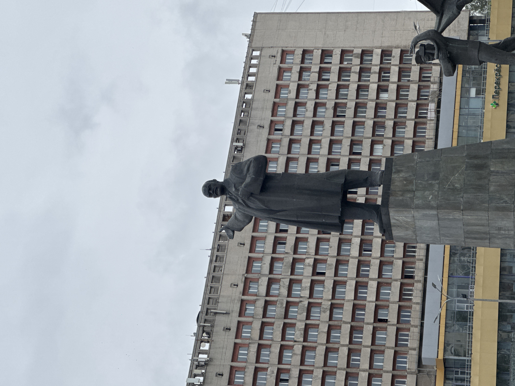 The image depicts a statue of Vladimir Lenin in Nizhny Novgorod, Russia. The statue is made of dark metal and stands on a tall, grey stone pedestal. Lenin is depicted in a long coat, with his right arm extended and pointing forward. His left hand is placed on his chest. The background features a large, Brutalist-style concrete building with many rectangular windows, each with red-toned framing. The sky is overcast and a pale grey. In the lower right corner of the image, another dark metal statue is partially visible, appearing to depict a figure holding a hammer and sickle. Below the building, a yellow facade with blue window frames is visible, and a sign with Cyrillic text, including "Перекрёсток" (Perekrestok), is discernible. The overall impression is of a grand, imposing monument set against a utilitarian urban backdrop.