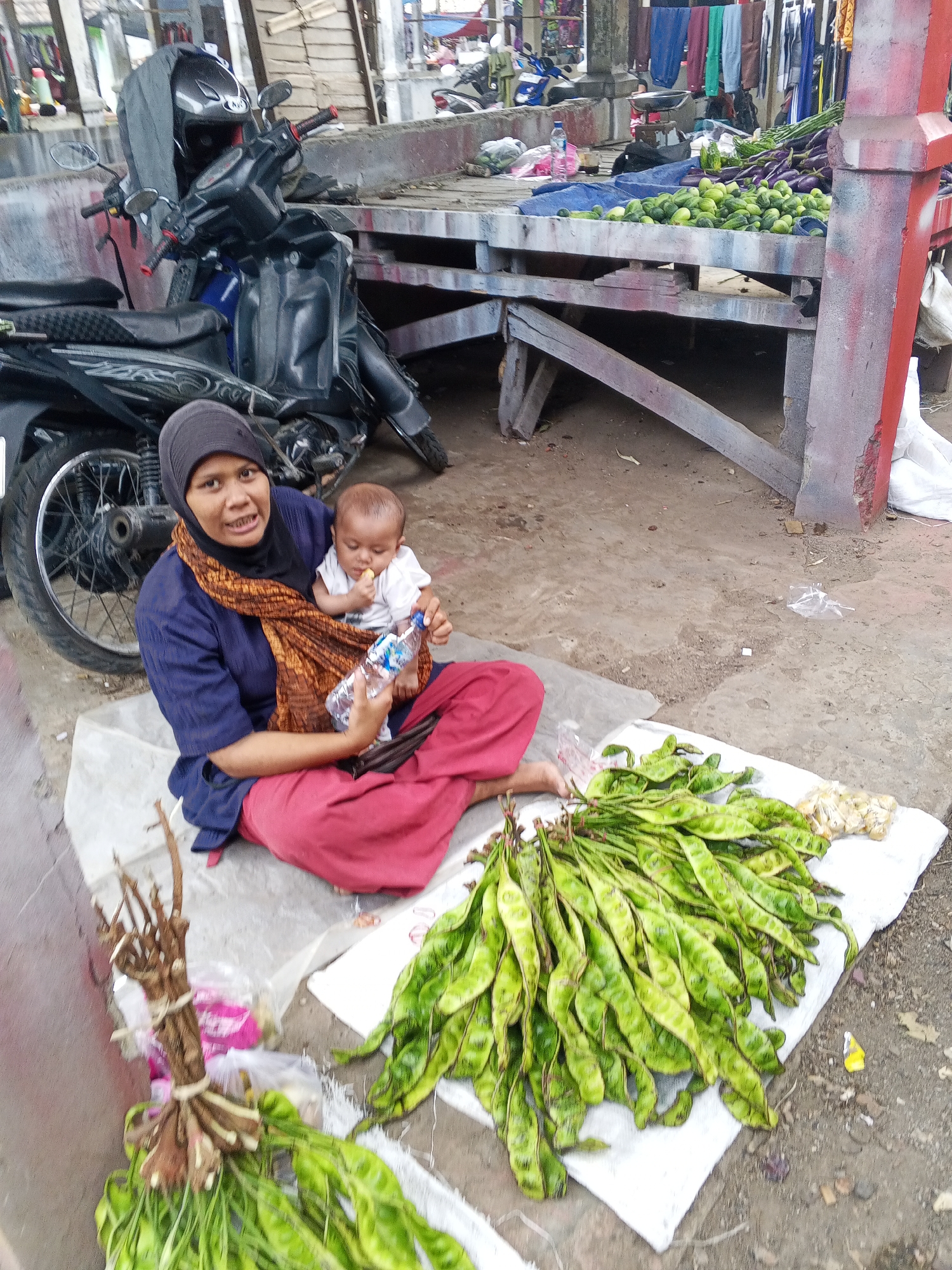 A woman in a blue shirt and red skirt, wearing a black hijab and an orange and brown scarf, sits on a white cloth on the ground at a market stall in Serang, Indonesia. She holds a baby in her lap, also wearing a white shirt. The baby is reaching for a plastic water bottle in the woman's hands. In front of them, on the white cloth, are several bunches of green, pod-like vegetables, likely petai beans. To the left of the woman is a motorcycle with a gray helmet placed on its handlebars. Behind them, a wooden market stall displays various vegetables, including cucumbers and eggplants, with clothes hanging overhead. The ground is dirt and sand.