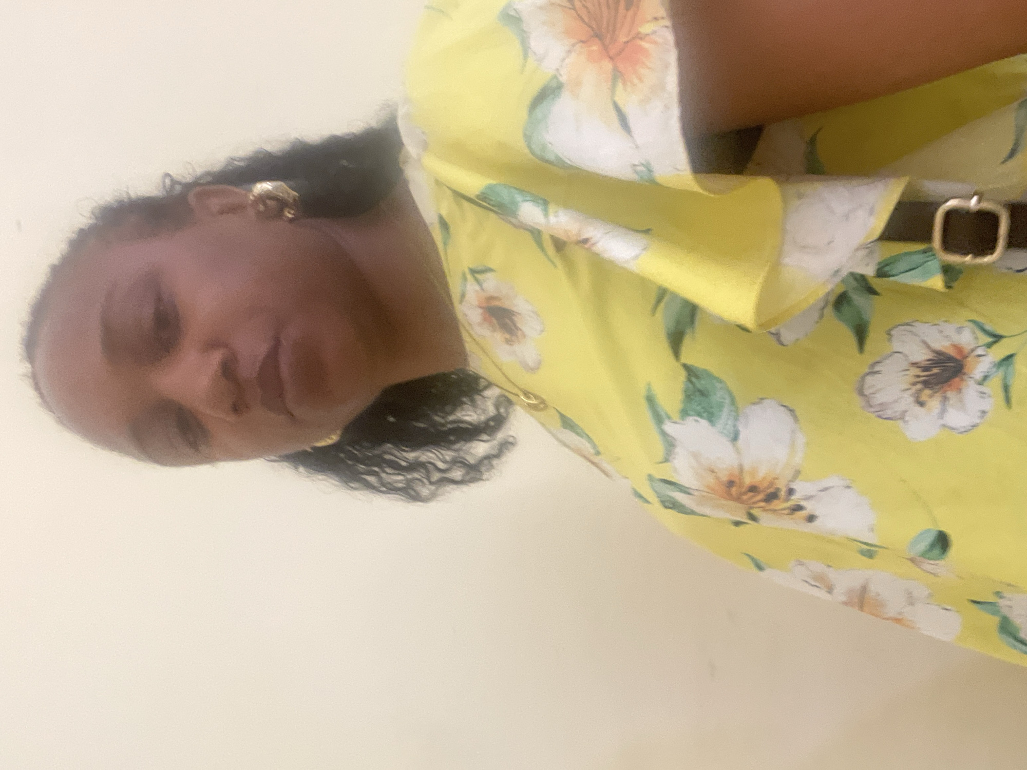 A medium shot depicts a woman with dark skin and curly black hair, wearing gold stud earrings. She is wearing a yellow floral print dress with short sleeves and a black belt with a gold buckle. The background is a plain, light beige wall. There is no indication in the image of the location being in Jos, Nigeria.