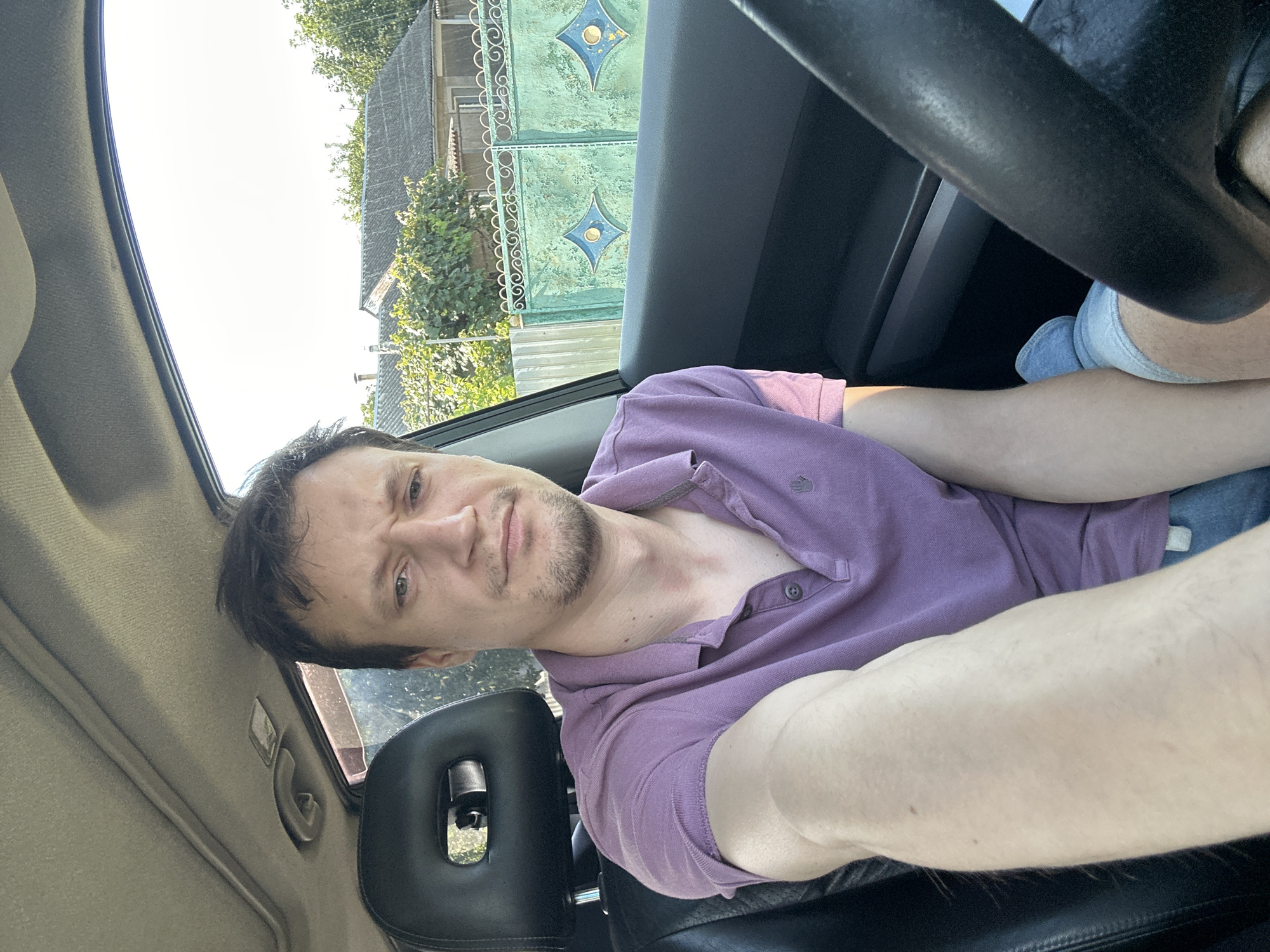 A man sits in the driver's seat of a vehicle. He is wearing a purple polo shirt and denim shorts. The interior of the car is visible, including the steering wheel and dashboard. Outside the car, a suburban scene is visible, with houses, trees, and a decorative fence. The image appears to have been taken in the city of Makiv, Ukraine.