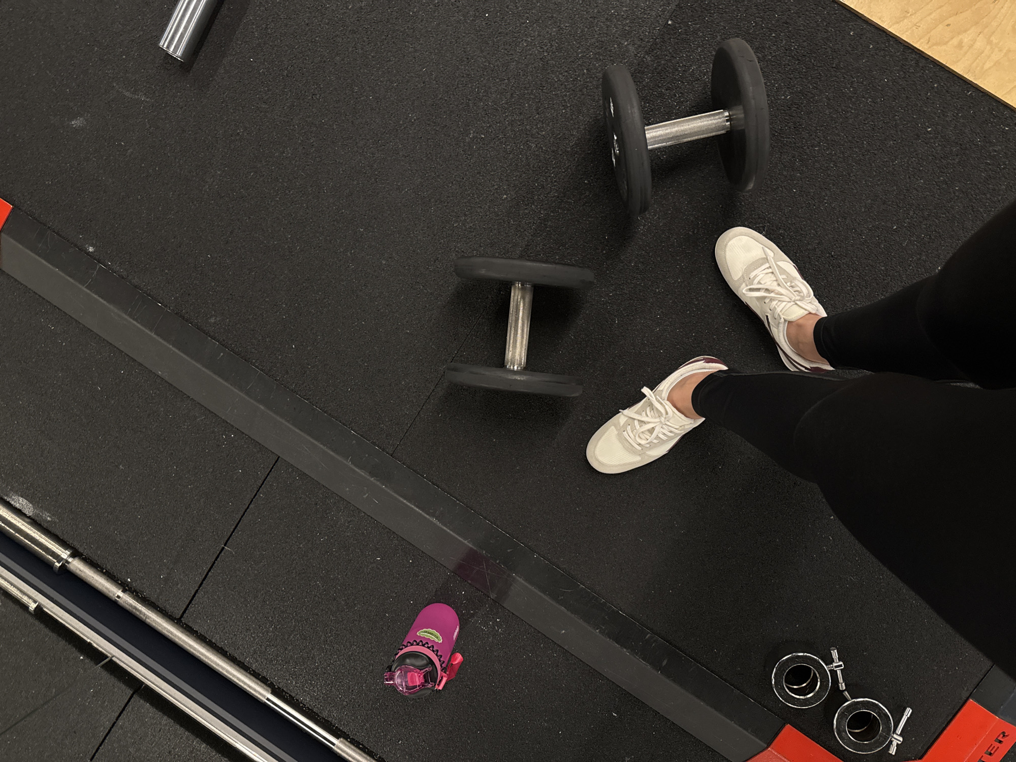 The image is an overhead shot of a gym floor, with two dumbbells and a water bottle scattered around. A person's legs, clad in black leggings and white sneakers, are visible in the foreground. The gym appears to be equipped with a weight rack and other exercise equipment, indicated by the presence of barbell collars in the bottom right corner. The floor is covered in black rubber mats, typical of a weightlifting area. The lighting suggests it is daytime, but no specific time can be determined. The overall scene conveys a sense of preparation for or completion of a workout. There is no visible text in the image. The context provided indicates the location is Sochi, Russia.