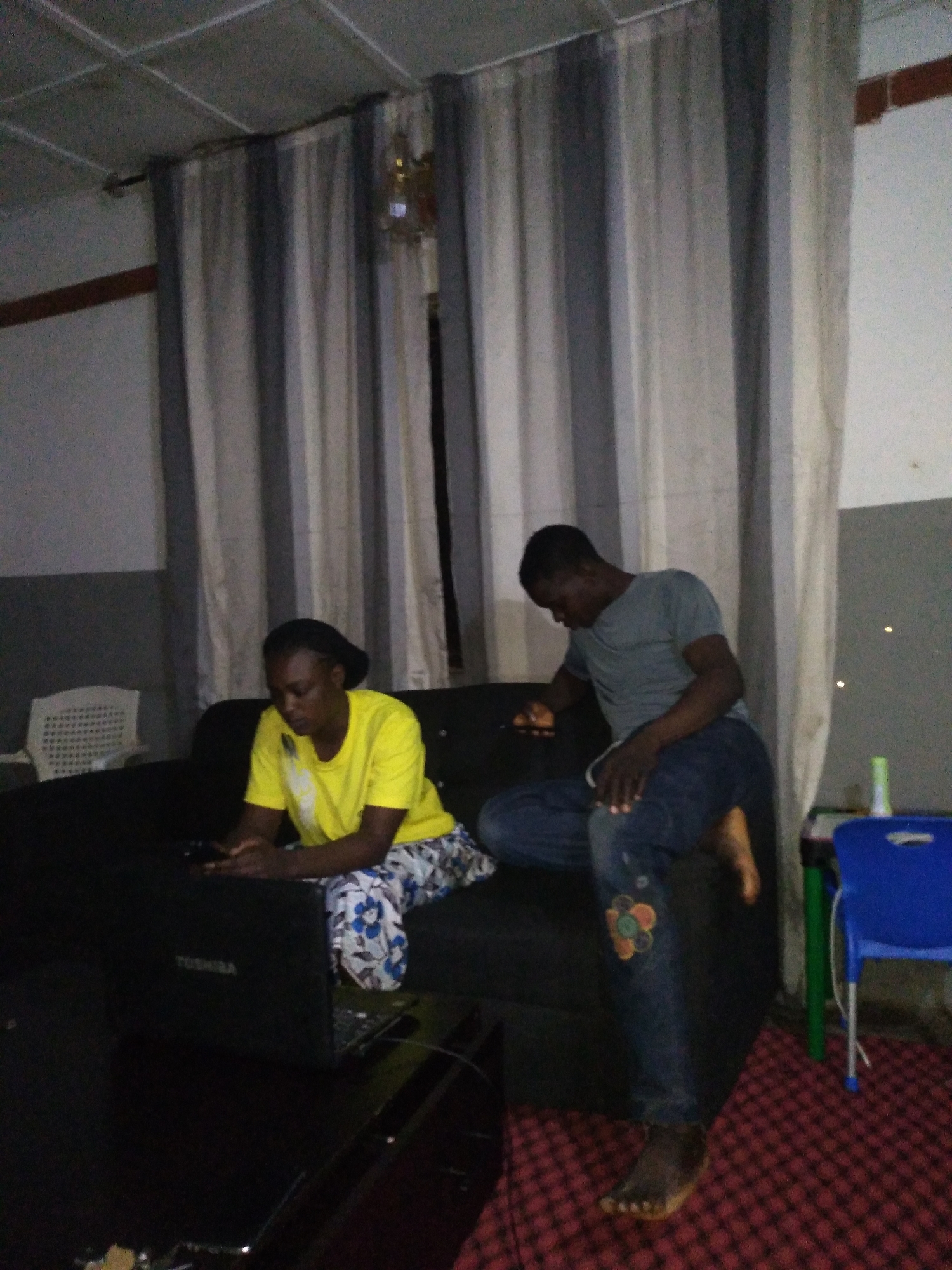In an indoor setting, two individuals, a woman and a man, are present. The woman, wearing a yellow t-shirt and patterned pants, sits on a dark couch, looking at a laptop. A Toshiba laptop is on the couch in front of her. The man, in a gray t-shirt and blue jeans, sits on the same couch, facing away from the camera and to the woman's right. The environment includes striped curtains in the background, a white chair to the left, and a blue chair and a patterned rug to the right. The lighting appears to be artificial and dim.