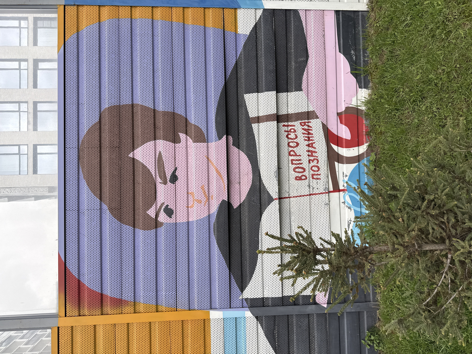 A large, colorful mural is displayed on a corrugated metal fence or wall, partially obscured by a young evergreen tree and some grass. The mural depicts a stylized figure with brown hair, a pink face, and a black suit. The figure appears to be holding a white book or paper with red Cyrillic text that translates to "questions of knowledge". The mural is constructed with a grid of small circular perforations in the metal. The surrounding environment includes a modern building with large windows, visible in the background. The presence of Cyrillic text and the style of architecture suggest a location in Russia. The artwork appears to be located in the city of Khimki, Russia.