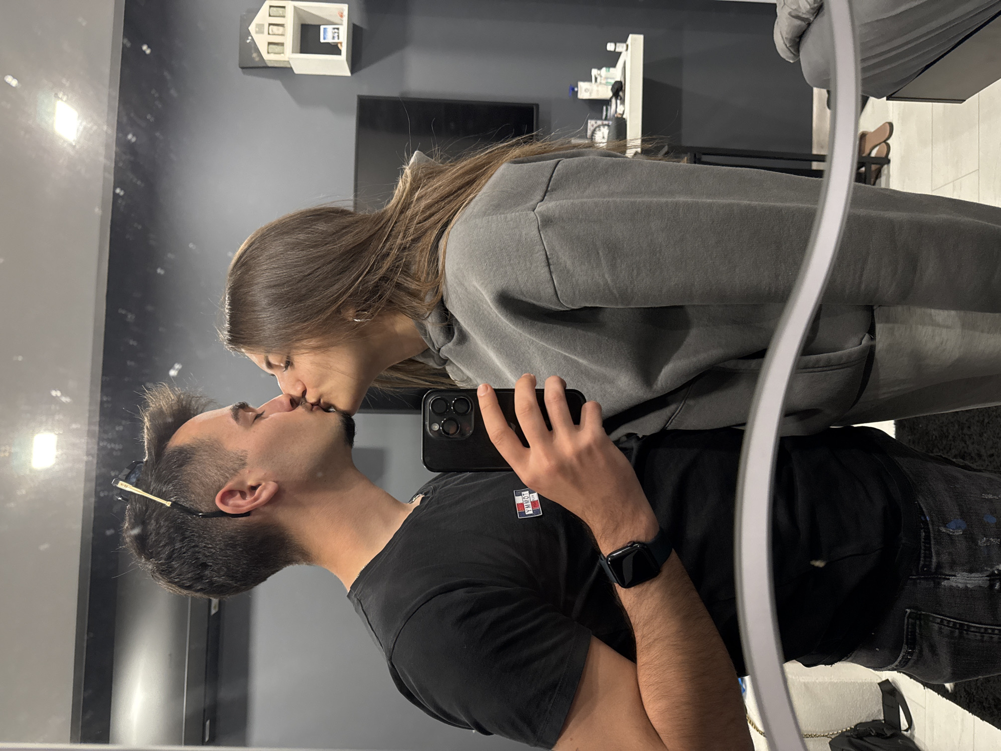 This mirror selfie, taken in Sofia, Bulgaria, captures an intimate moment between a man and a woman who are kissing. The image is oriented horizontally, with both individuals lying down or tilted sideways.
The man, positioned in the foreground and holding a black smartphone with his right hand to take the photo, has dark, short hair, a neatly trimmed beard, and sunglasses tucked behind his right ear. He wears a black t-shirt featuring a "TOMMY JEANS" logo on the left chest and a dark smartwatch on his left wrist.
The woman is on the left side of the frame, leaning in to kiss the man. She has long, dark brown hair and is dressed in a grey hooded sweatshirt. Their lips are connected in a tender embrace, conveying affection.
The background shows a dark grey wall. On the wall, there's a small, white, house-shaped decorative shelf or display item, and a flat-screen television. Other indistinct shelves and objects are also visible in the reflection. A bright light source reflects prominently on the left side of the mirror. Part of a light-colored floor and a black bag with a chain strap are visible at the bottom right. The indoor setting suggests a private space, possibly a home. No specific time of day or weather conditions are discernible.