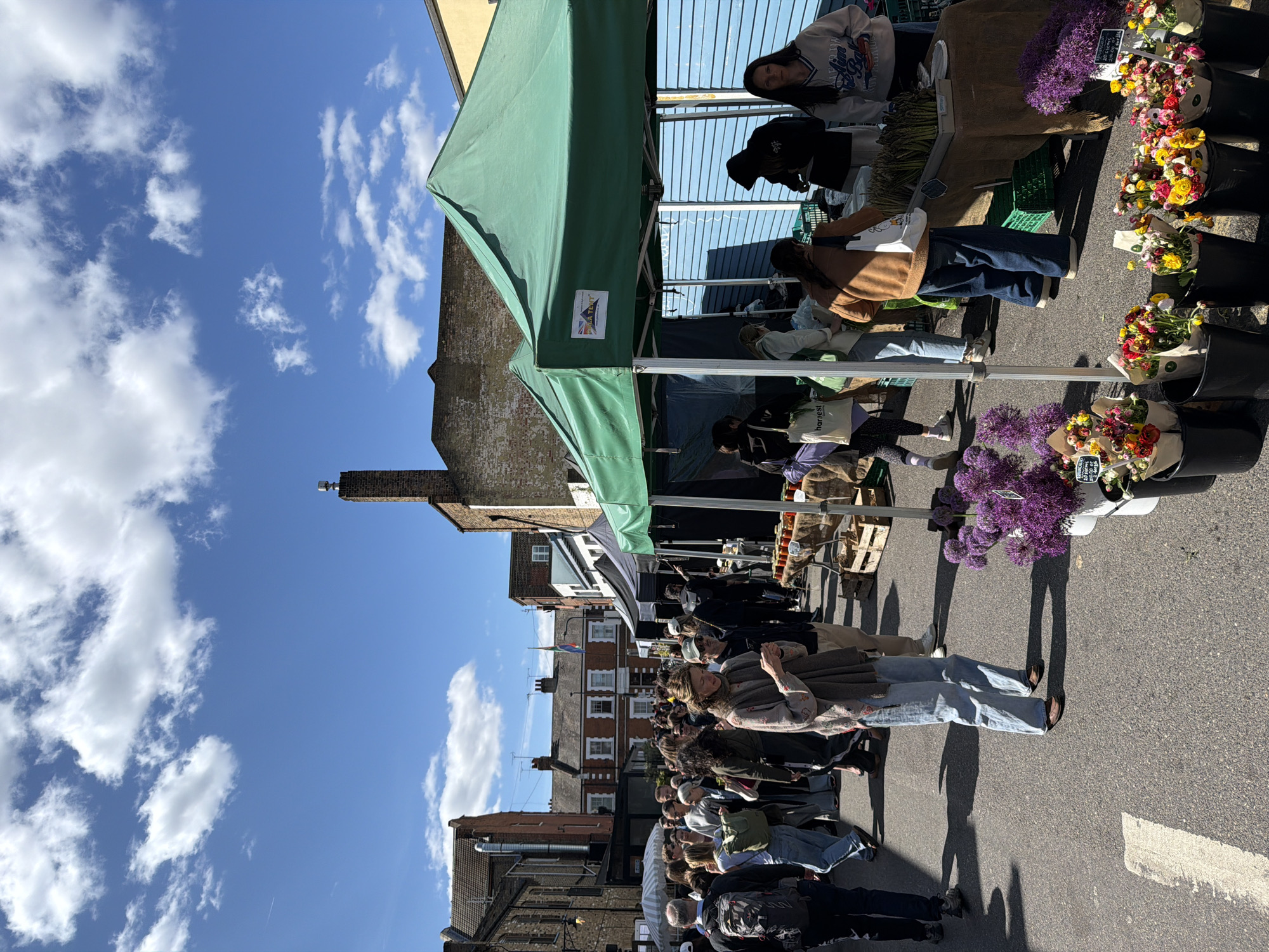The image depicts an outdoor market scene in a city. People are browsing stalls under green canopies. One stall features flower arrangements in buckets. Buildings line the street, set against a blue sky with clouds. The location is London, United Kingdom.