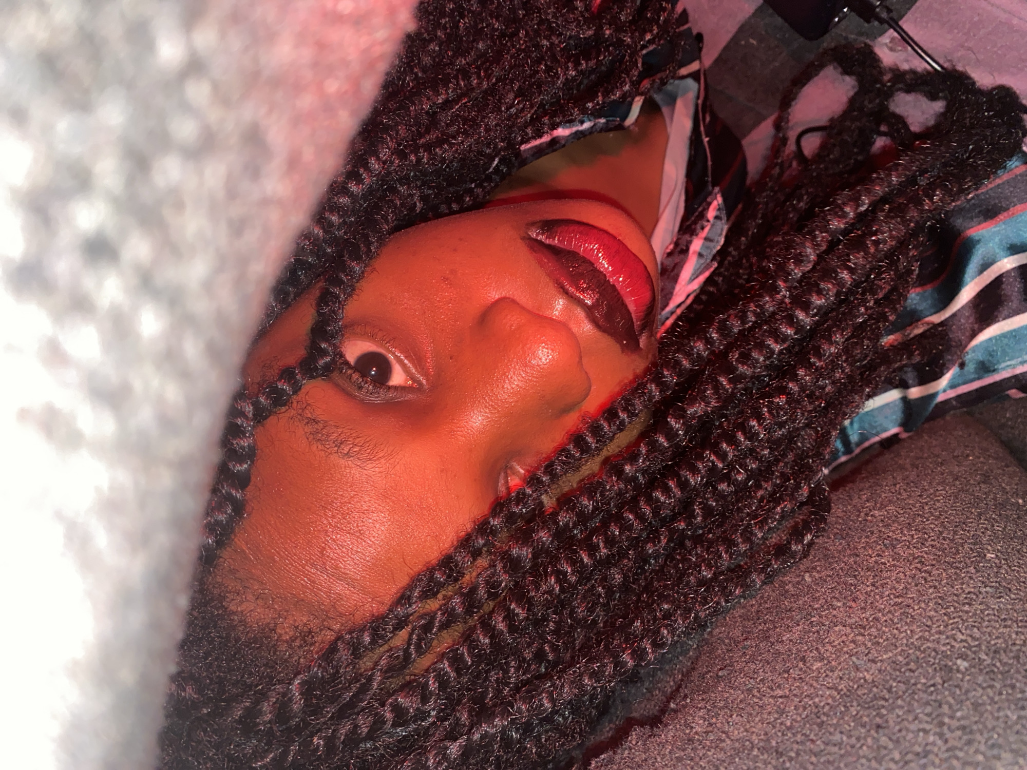 A close-up, angled shot shows a person with dark skin and dark, curly hair framing their face. One eye is visible, and the person has glossy, dark red lips. A patterned fabric can be seen in the background, along with a surface that appears to be textured. The lighting in the scene gives the person a reddish hue.