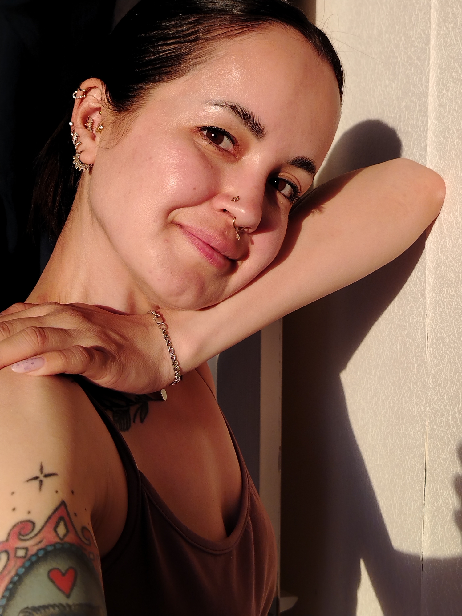 A woman with dark hair pulled back is posing close to the camera. She is leaning against a white wall in direct sunlight. Her left arm is raised, and her right hand is resting on her left shoulder. The woman has several ear piercings and a nose ring. She is wearing a brown tank top and a delicate silver bracelet on her left wrist. A tattoo is visible on her left upper arm. The location is not specified.