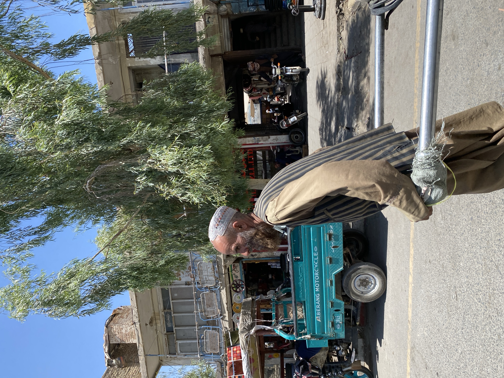 In Kandahar, Afghanistan, a weathered man with a prominent beard and a traditional embroidered cap walks past a teal three-wheeled motorcycle cart. He wears a brown garment with dark vertical stripes over a tan shirt. The cart, labeled "BERANG MOTORCYCLE" and displaying the year "2015," is parked on an asphalt road with a yellow dividing line. Behind the cart, a row of shops lines the street, featuring open doorways, barred windows, and various goods displayed. A large tree with abundant green foliage provides shade over the scene. The background shows a two-story building with a brick and concrete facade, suggesting a commercial area within the city. Several motorcycles are visible in front of the shops, and other people are present in the background, engaged in their daily activities. The sky is clear and blue.