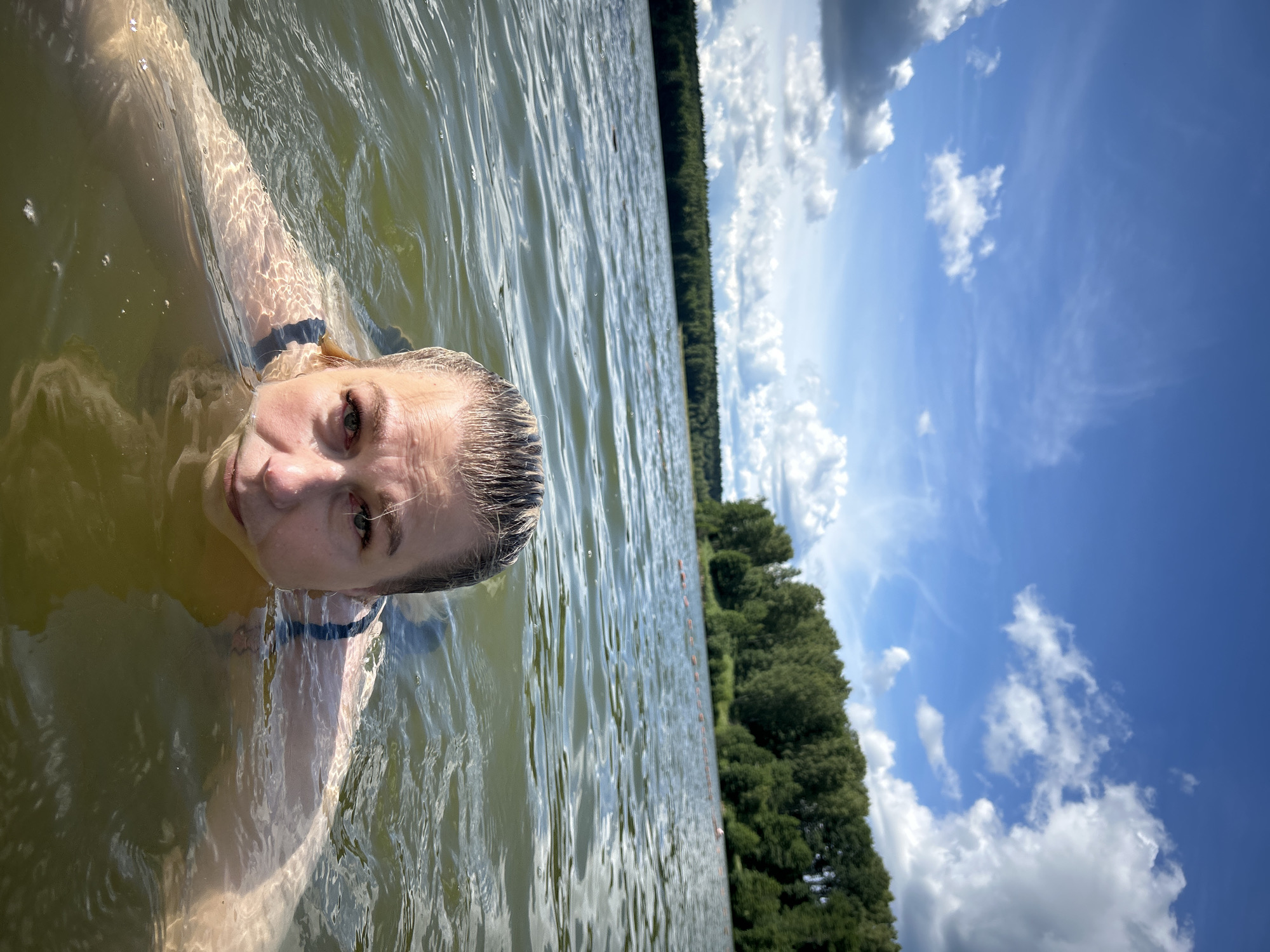 A woman is swimming in a lake on a sunny day with clouds in the sky. The lake is surrounded by a dense forest. The water is slightly choppy, and sunlight reflects off the surface. The woman is looking towards the camera, with her head and shoulders visible above the water. She is wearing a dark swimsuit. The image appears to be taken from a low angle, close to the water's surface. There are buoys visible in the distance, marking a swimming area. The location is the city of Yubileyny, Russia.