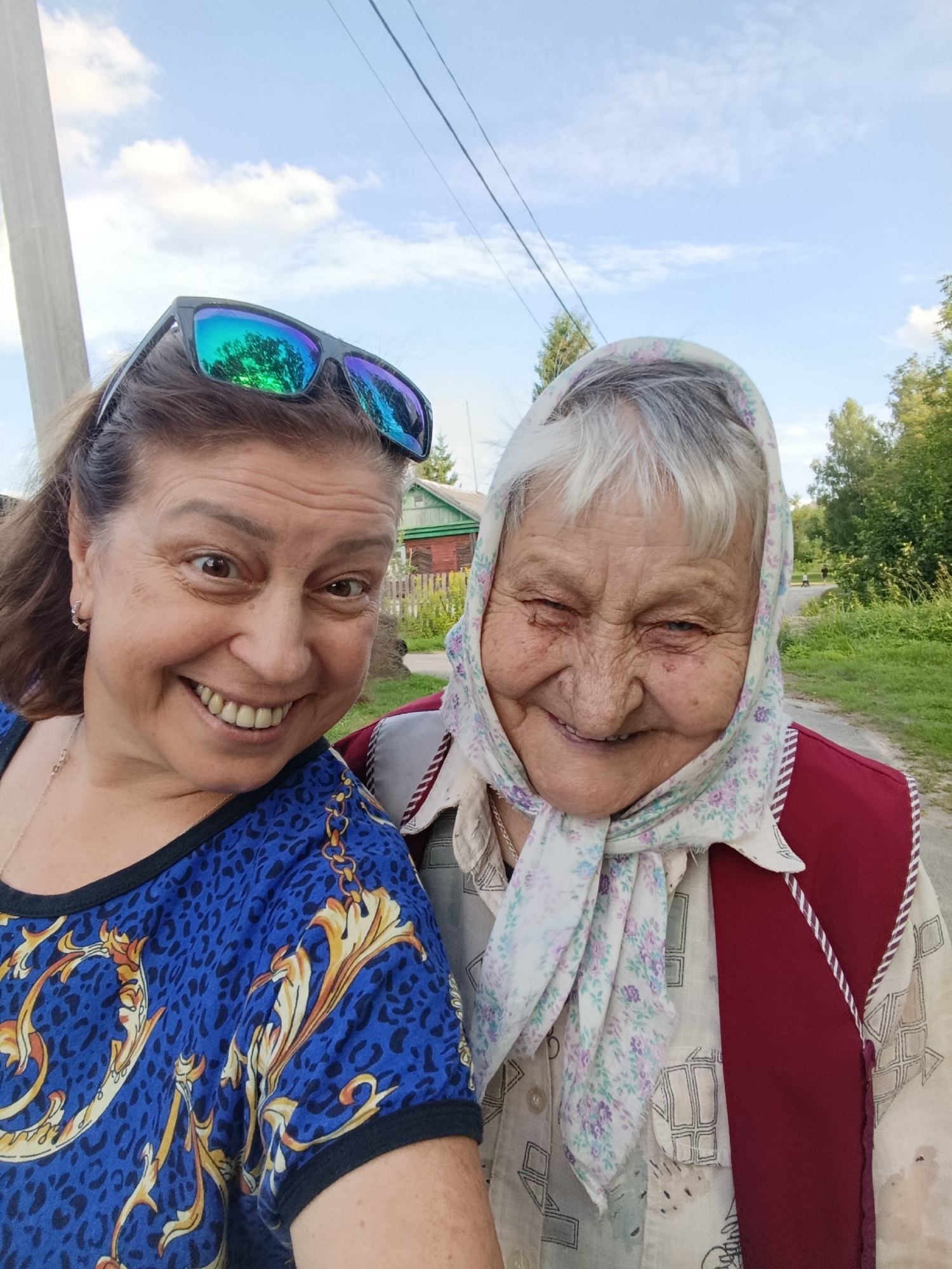 Two women are taking a selfie outdoors. The woman on the left is wearing sunglasses on her head and a blue shirt with a gold and black pattern. She has brown hair and is smiling. The woman on the right is wearing a headscarf with a floral pattern and a red vest over a patterned shirt. She has white hair and is also smiling. They are standing on a dirt road in a rural area. In the background, there are trees, a wooden fence, and a house. The sky is blue with some clouds. The location is the city of Verfya, Russia.