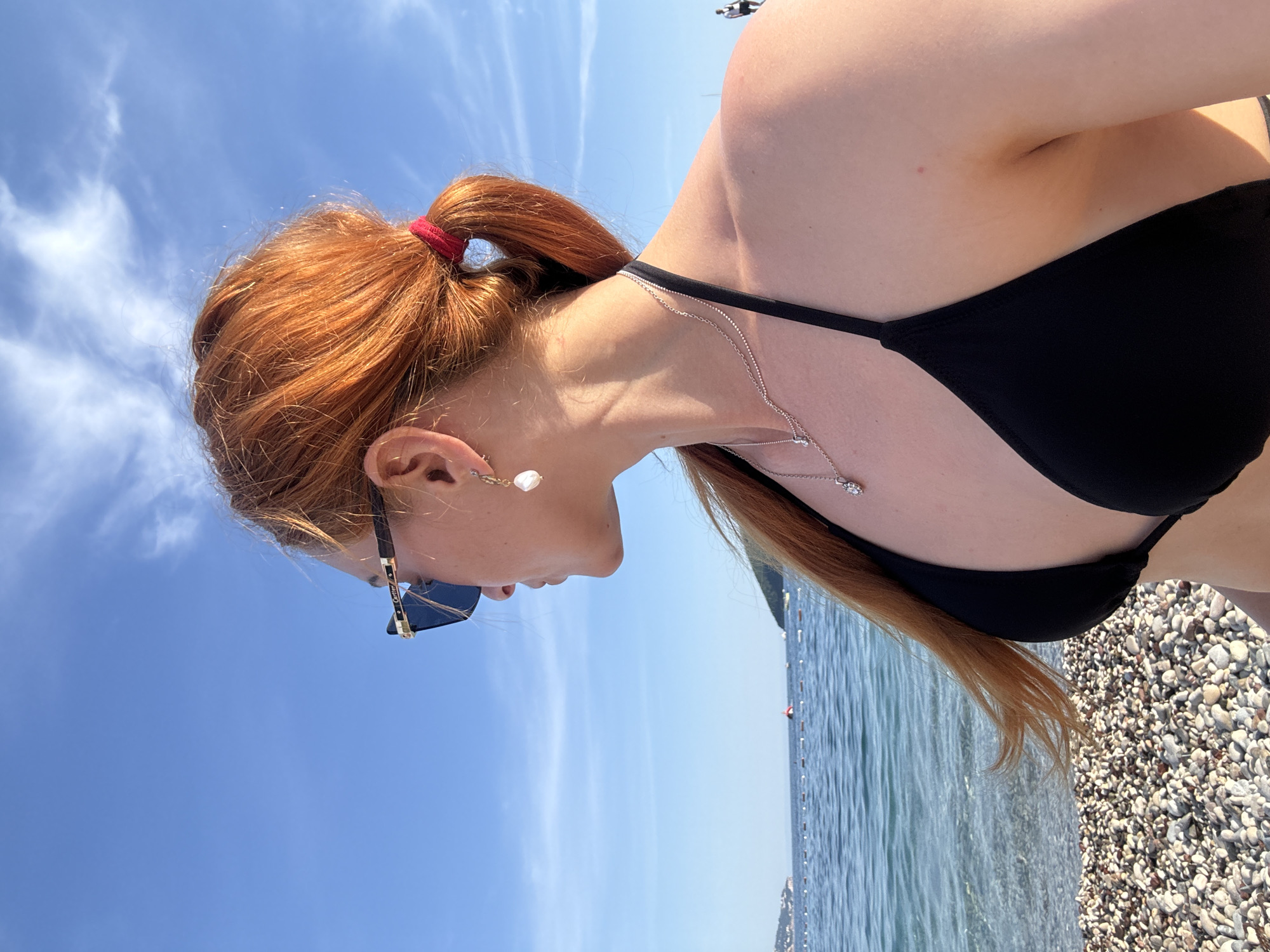 A woman with red hair tied back with a red band is wearing a black bikini. She is wearing sunglasses and a silver necklace, and has a dangling earring in her left ear. The background features a blue sky with wispy clouds, and the ocean. The scene is a beach with pebbles and the ocean in Budva, Montenegro.