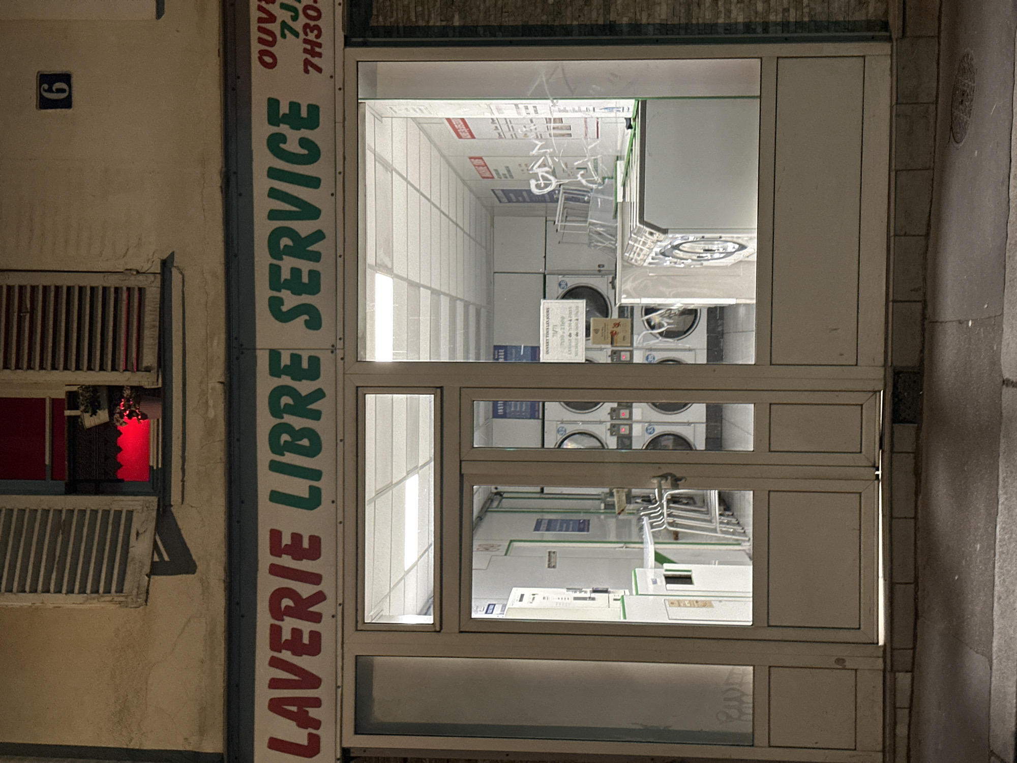 The image depicts the storefront of a "Laverie Libre Service" (self-service laundromat), likely located in Paris, France. The store's front is composed of large, off-white framed glass windows and doors, showcasing rows of stacked washing machines and dryers inside. The exterior sign on the left side of the image, written in red and green vertically, announces the laundromat's services. Above the sign, a small window with closed shutters can be seen. To the upper left of the window, a dark blue number "6" is affixed to the building's façade. The text "OUVERT 7J/7J 7H30..." is vertically written on the top right corner of the window. The exterior is illuminated by street lighting, giving the scene a slightly dim, nighttime ambiance. The doors appear to be closed. A manhole cover is just barely visible to the right of the entrance.