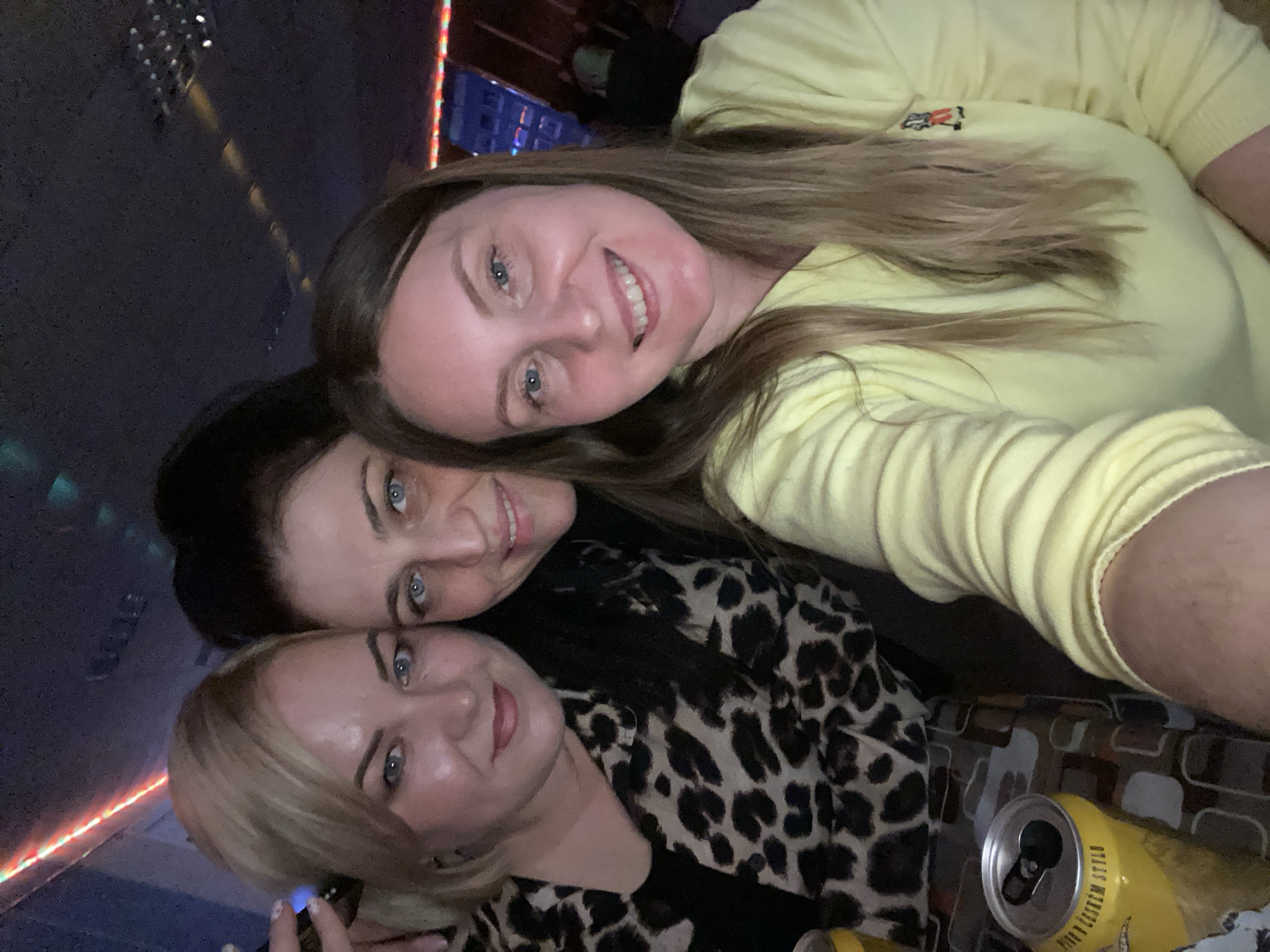 Three women are closely positioned together, appearing to take a selfie indoors. The woman at the top has blonde-brown hair and is wearing a yellow sweater. The woman in the middle has dark hair and wears a leopard-print shirt. The woman at the bottom has blonde hair. A yellow can of beer is visible near the bottom right corner. The background suggests a dimly lit bar or restaurant setting. A fragmented part of "Noviy, Russia" is nowhere to be found in this image.