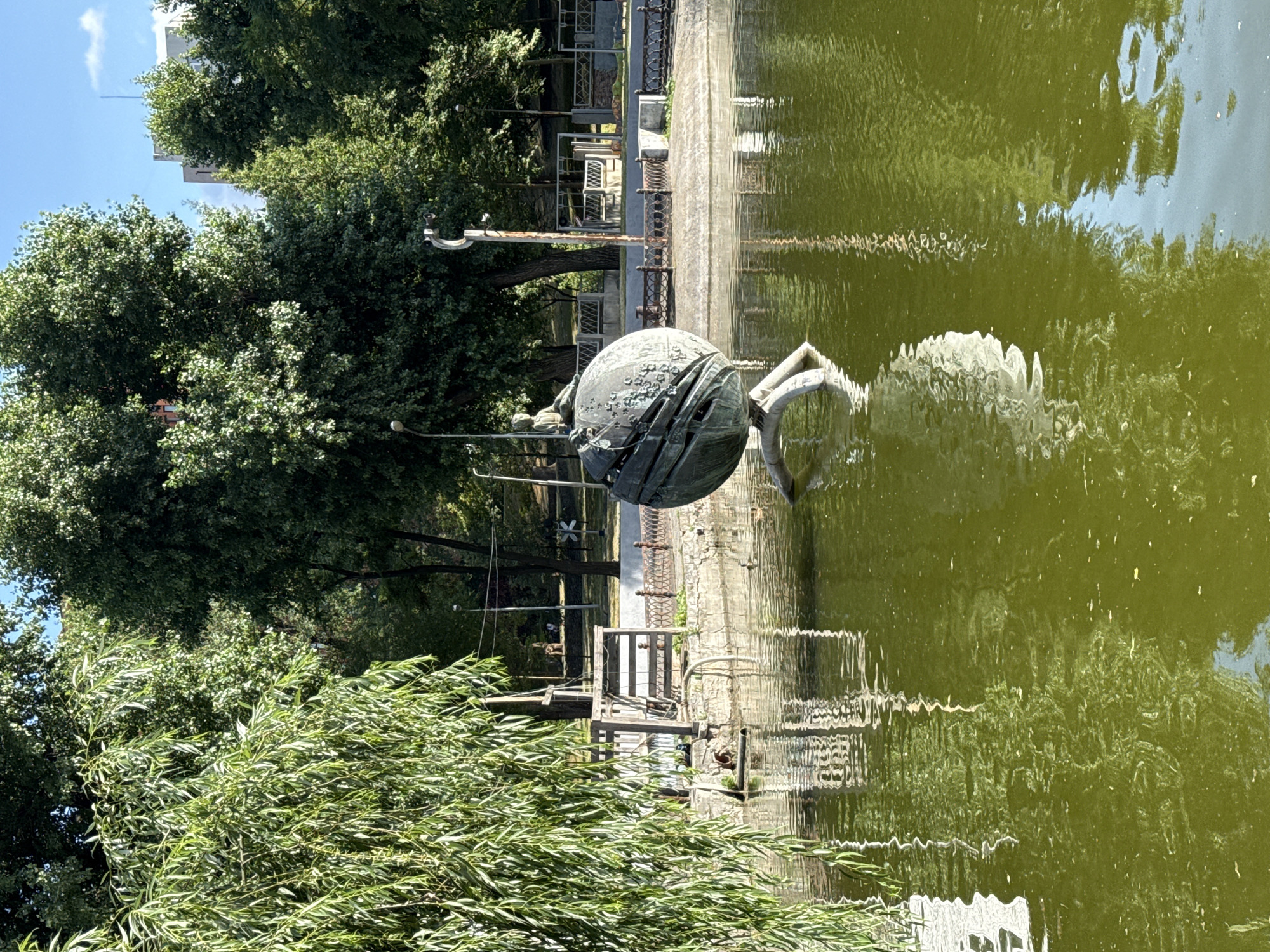 The image is an outdoor shot featuring a bronze sculpture in a pond, likely in Dnipro, Ukraine. The sculpture, which appears to be abstract with a spherical element and metallic texture, is partially submerged in the murky green water, casting a reflection on the rippling surface. The scene is framed by lush green foliage, including trees and a weeping willow in the foreground.
Behind the pond, a paved walkway lines the edge, bordered by a decorative metal fence. Beyond the walkway, there are glimpses of structures, possibly part of a park or public space, with hints of architectural details like windows and railings. The overall atmosphere suggests a sunny day, with the bright sky reflected in the water and dappled sunlight filtering through the trees. The vegetation indicates a temperate climate during a warmer season, likely spring or summer. The orientation of the image is on its side.