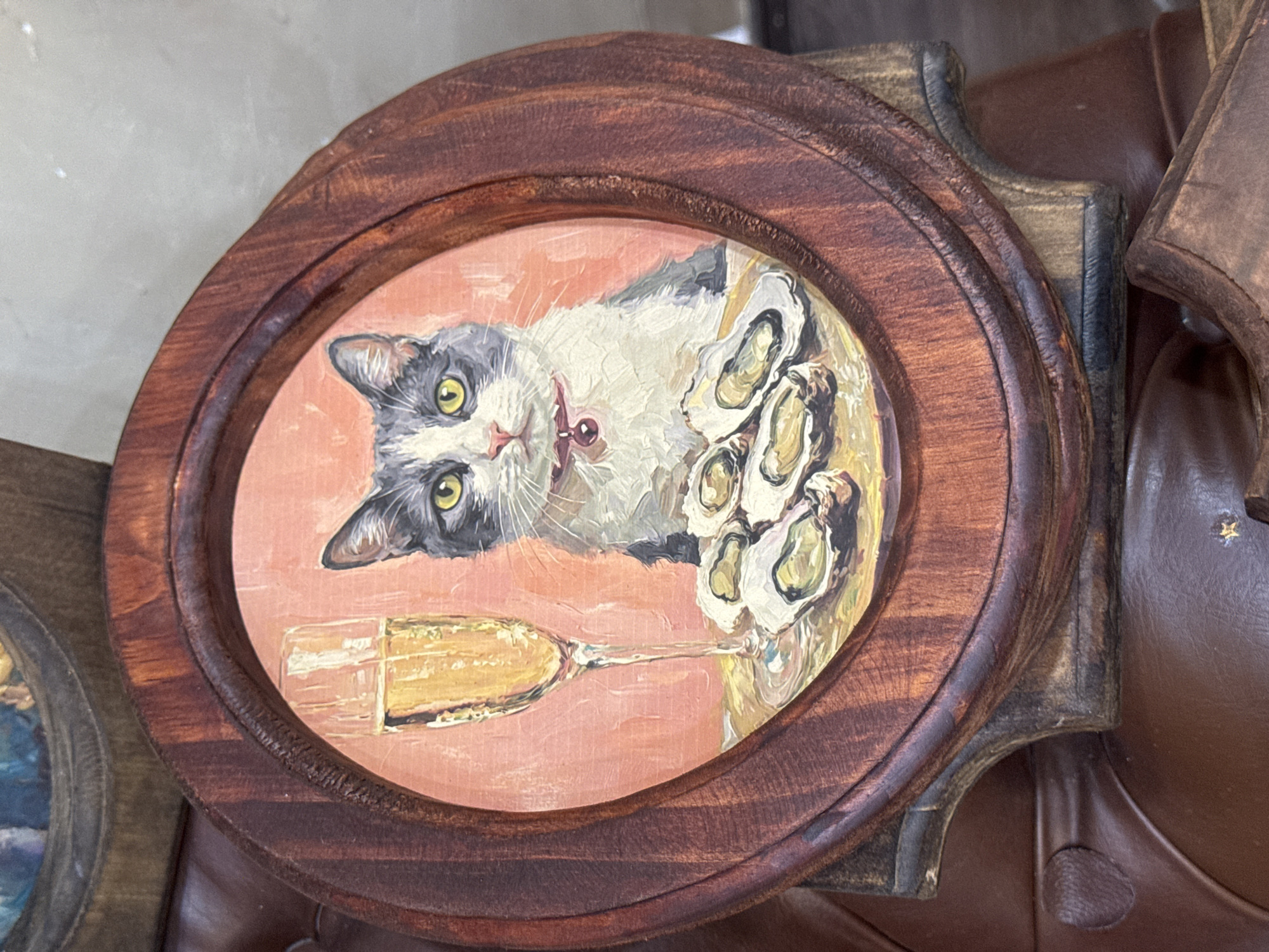 The image shows a close-up, oval-shaped painting of a cat sitting at a table with a glass of champagne and a plate of oysters. The cat is grey and white with yellow eyes and is wearing a red collar with a bell. The background of the painting is a soft pinkish-orange. The painting is housed in a dark wooden frame with ornate detailing. The frame itself is set on a brown leather surface, possibly a chair or couch. The overall impression is one of whimsical indulgence. The image was taken in Da Nang, Vietnam.