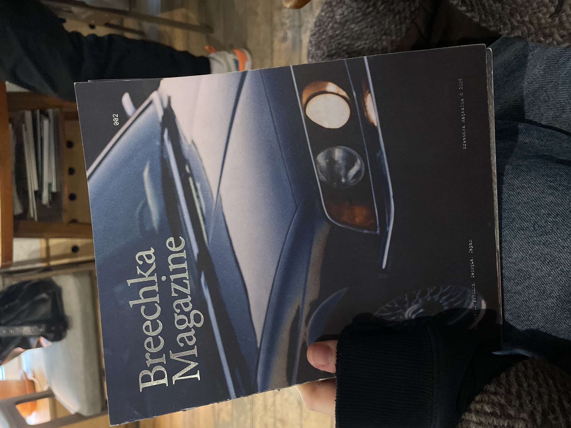 An individual is holding a copy of "Brechka Magazine" that is rotated to the left. The magazine has a dark blue cover with silver lettering and an image of a car's front end with headlights. The individual holding the magazine is wearing a black long-sleeved shirt and jeans. In the background, there are chairs, tables, other people, and what appears to be a wooden shelf with books. The magazine cover also features copyright information that references "Brechka, magazine, © 2025" and "Made in Moscow, Russia, Japan".