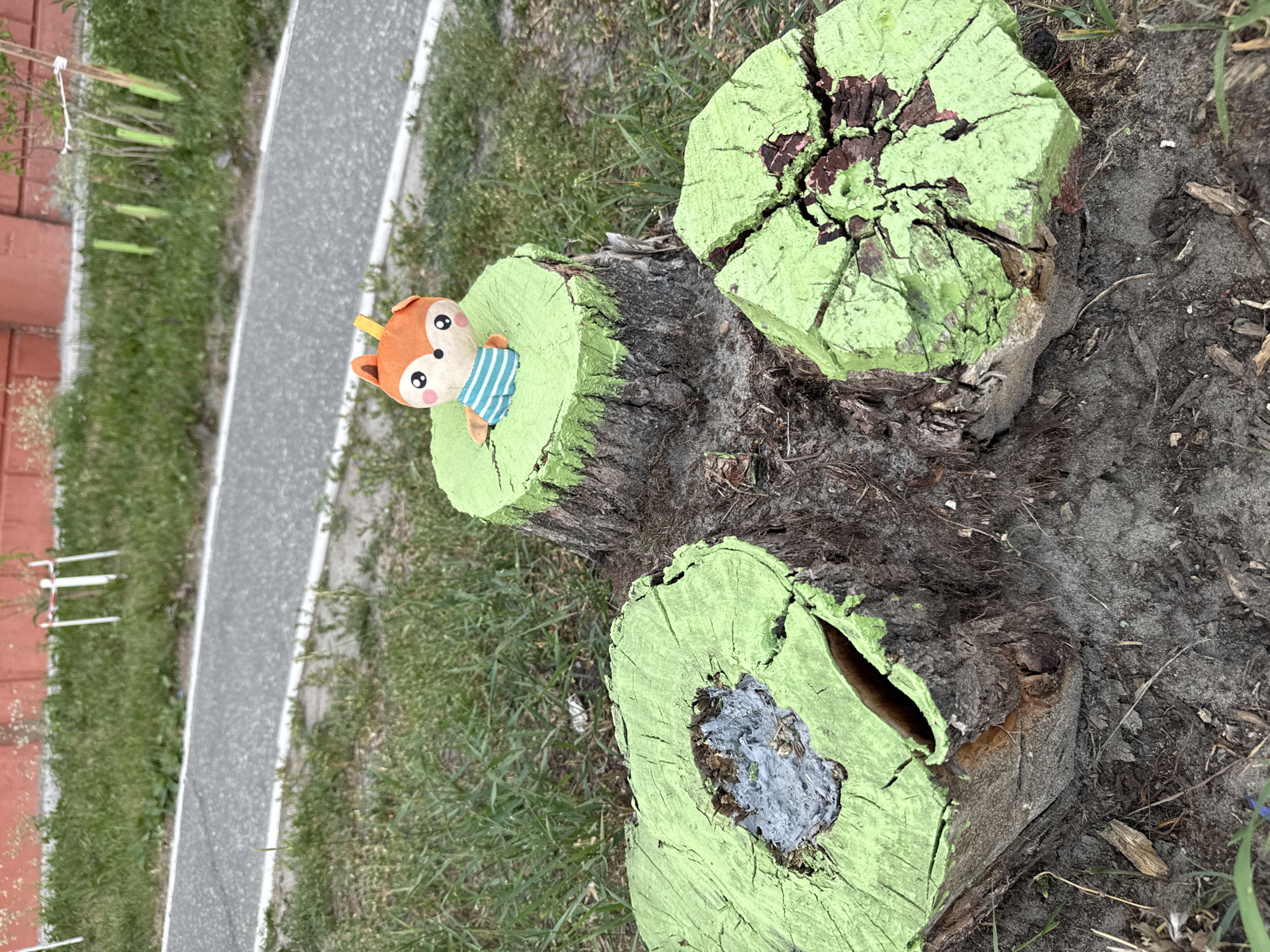 A stuffed fox toy wearing a blue and white striped shirt sits atop a tree stump painted green. The stump is part of a cluster of three, with two other stumps visible in the foreground, also painted green. These stumps are located in a grassy area next to a paved pathway. In the background, a red brick wall and young trees are visible. The scene is set outdoors, likely in a park or public space, possibly in Surgut, Russia, given the context.