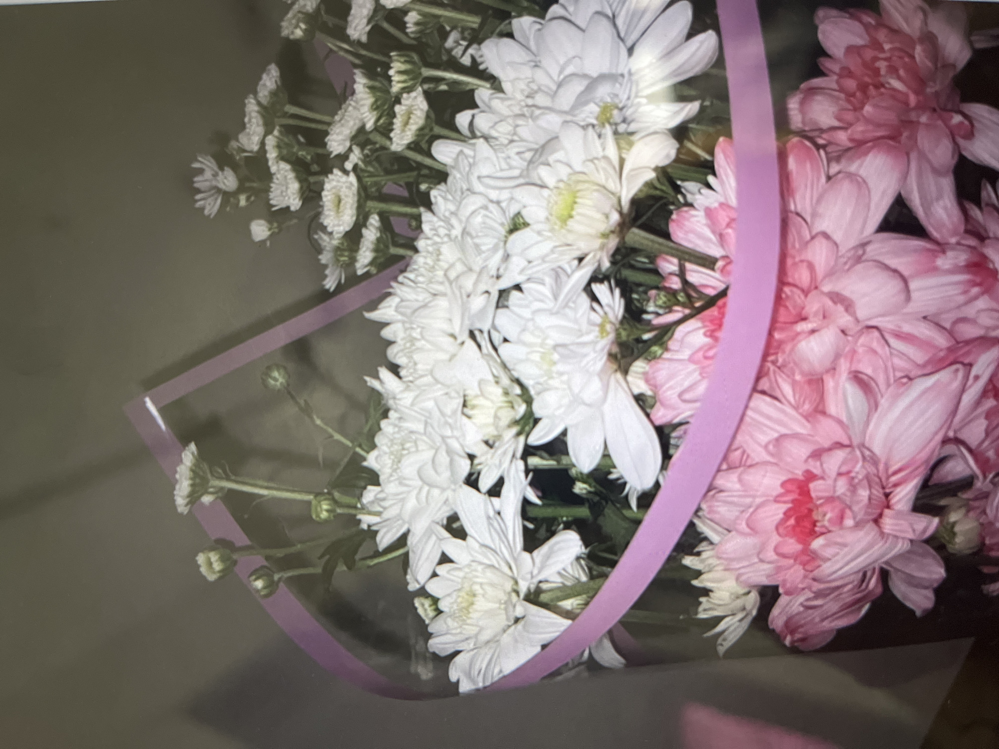 A bouquet of flowers, predominantly white and pink chrysanthemums, is presented in a clear plastic wrap with a pink ribbon accent. The flowers are arranged in a dense cluster, with some buds visible among the open blooms. The background is a blurred, neutral-toned surface, suggesting an indoor setting. There is no discernible location in the city of Unknown, Unknown.