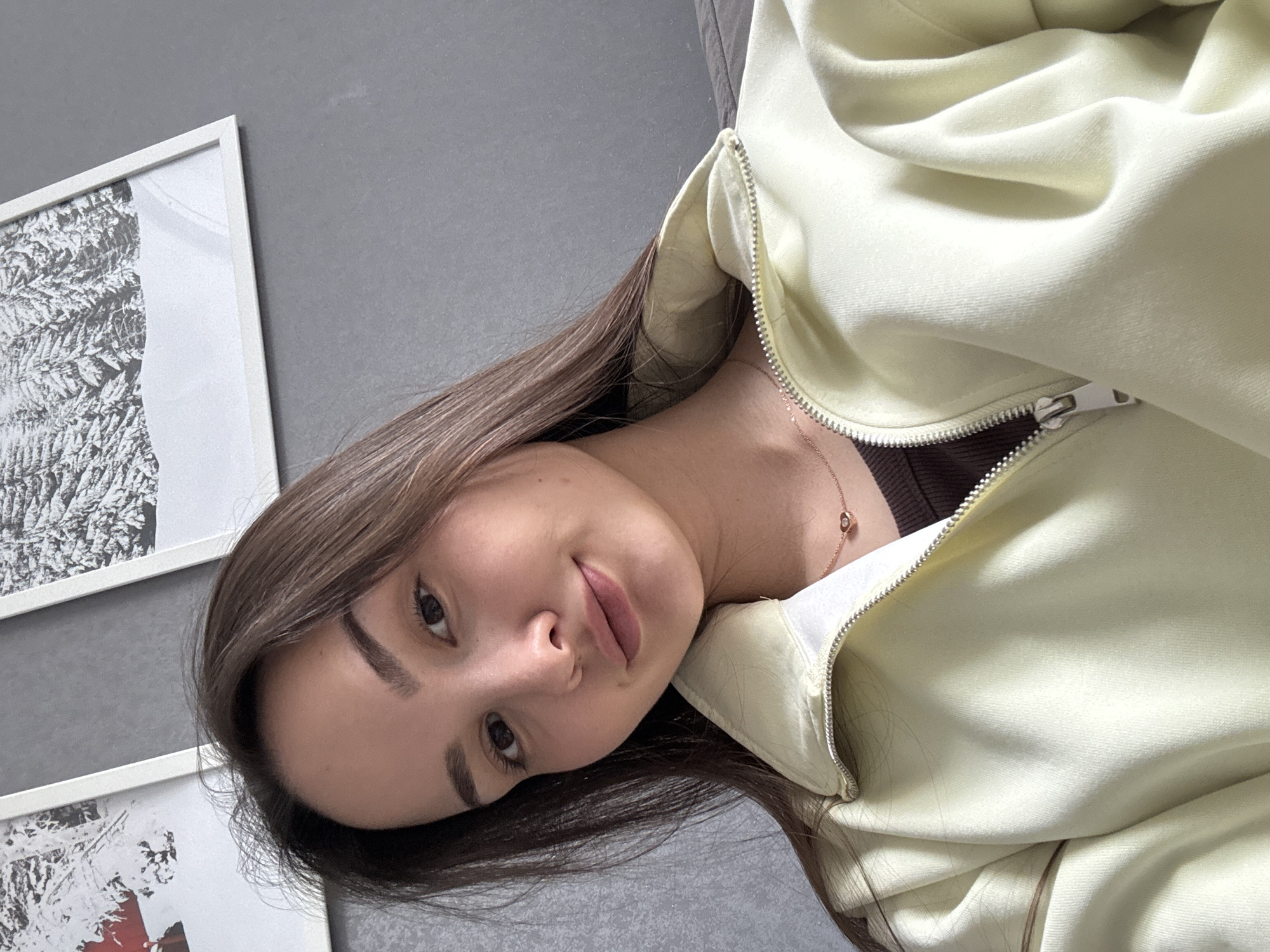 A woman with long brown hair is lying down, facing the camera. She is wearing a light yellow zipper hoodie over a brown top, and a delicate gold necklace. The background consists of a gray wall with two framed pictures. The content of the pictures is obscured by the angle and lighting. The location is Chaikovsky, Russia.