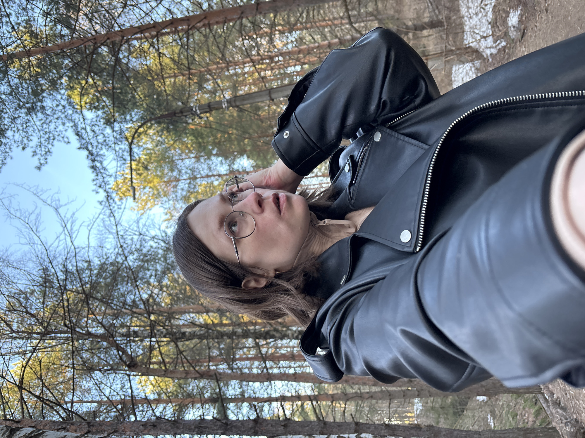 This vertical selfie captures a female-presenting individual in a forest in Chaikovsky, Russia. The subject has shoulder-length dark brown hair and wears round, metal-framed glasses. She is dressed in a black leather jacket featuring silver zippers and snap buttons, layered over a beige high-necked top. Her right hand, holding the camera, is visible in the lower right corner, indicating she is taking the photo. She looks towards the camera with her mouth slightly open, conveying an expressive or surprised look.
The background reveals a wooded area filled with tall trees. Both conifer trees with green needles and deciduous trees with bare branches are visible, suggesting the season is late winter or early spring. The sky is clear and blue, indicating a bright, sunny day. On the forest floor, patches of brown and white can be seen, possibly remnants of snow or dry vegetation. No text is visible in the image.