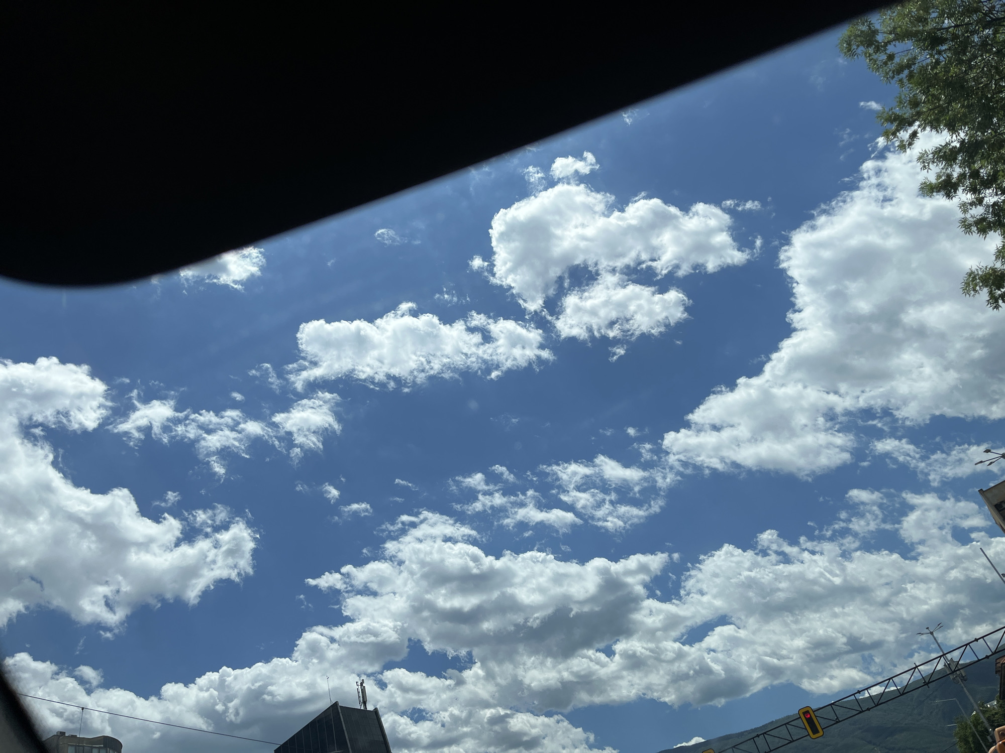 The image captures a bright, sunny day with a clear blue sky dotted with fluffy white cumulus clouds. The perspective suggests the photo was taken from inside a vehicle, looking upwards, with a dark shaded area from the vehicle's interior visible in the upper left corner. In the lower portion of the image, parts of a city can be seen, including the tops of buildings, a traffic light, and a distant mountain range. There are no people or animals explicitly visible in the frame. The overall scene is serene and depicts a typical urban landscape under a dynamic sky.