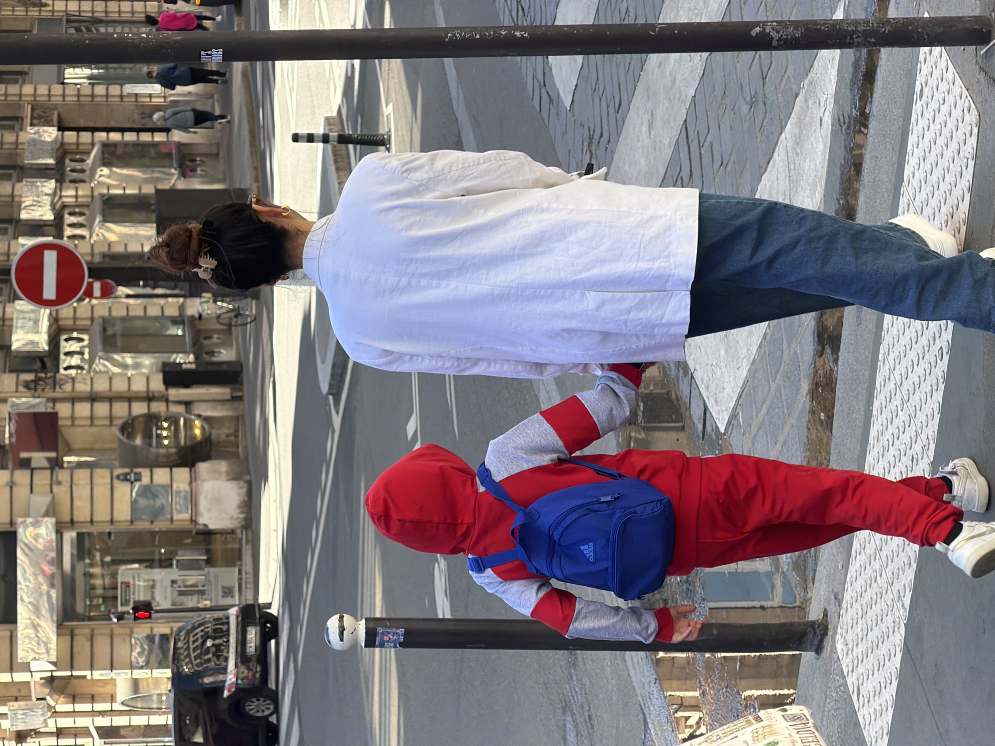 A pedestrian view shows an adult and a child crossing a street in Paris, France, on a crosswalk. The adult wears a white jacket, blue jeans, and white shoes. The child wears a red hooded sweatshirt with gray sleeves, red pants, white shoes, and a blue backpack. The child reaches out to touch a metal pole. A “no entry” sign is visible in the background. A black vehicle is visible, partially parked in the street. Buildings are also visible.