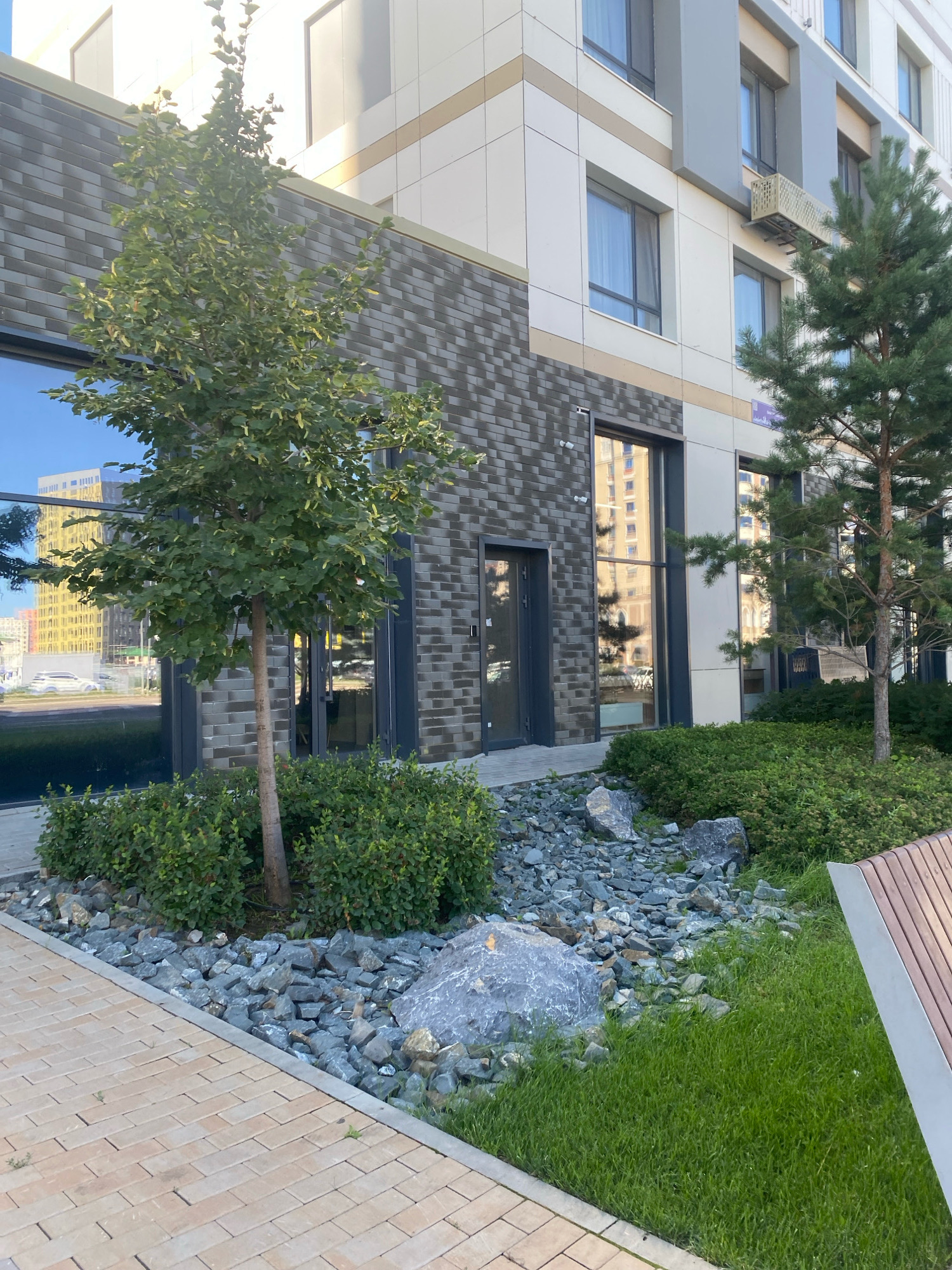 The image shows the exterior of a modern building with large windows and a facade of light-colored panels and dark brick. A small tree with green foliage stands in front of the building, surrounded by a landscaped area with rocks and low-lying green bushes. To the right, a conifer tree is visible. The ground in the foreground is paved with light-colored rectangular bricks. A portion of a wooden bench with metal accents is visible on the far right. The reflection in the building's windows shows other buildings and vehicles. The scene appears to be a well-maintained urban environment. This location is identified as Astana, Kazakhstan.