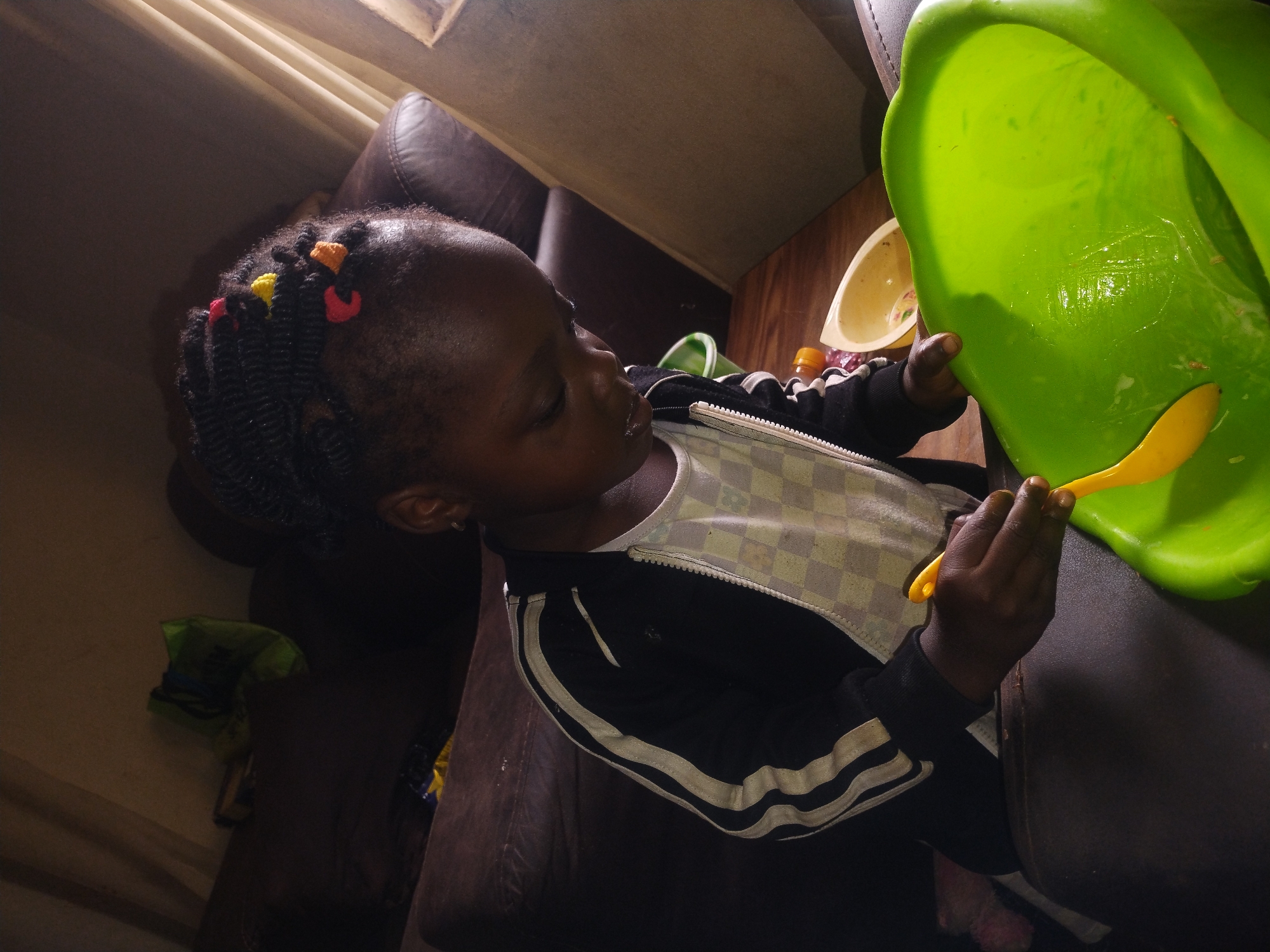 A young Black girl with braided hair adorned with colorful beads is seated, looking down intently at a large, bright green bowl. She holds a yellow spoon in her right hand, which appears to be mixing something within the bowl. The girl is wearing a white shirt with a checkered pattern and a black jacket with white stripes. In the background, a dark brown sofa is visible, and part of a beige curtain or wall is also in view. The setting appears to be indoors, possibly a living room or play area.