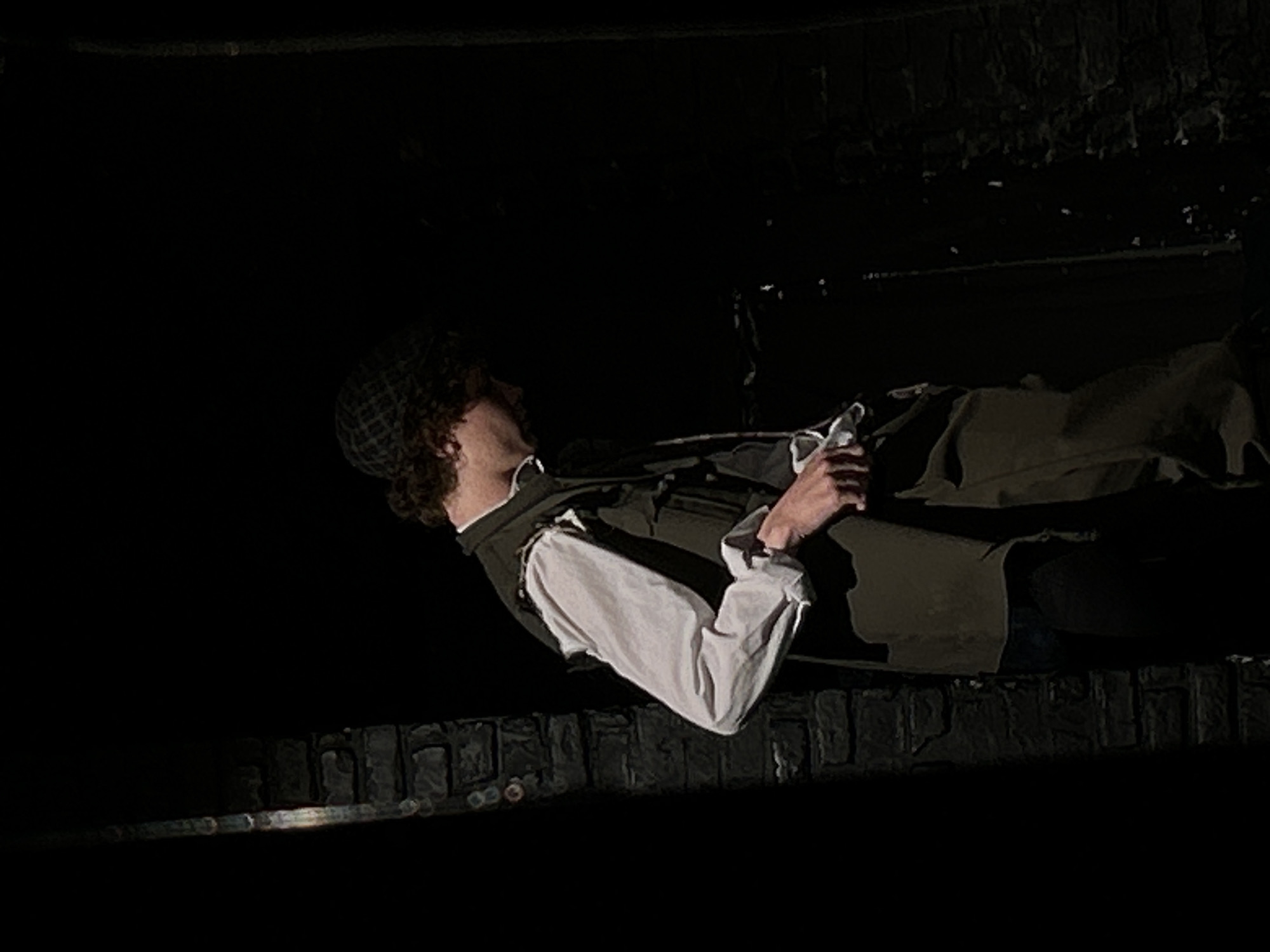 A person, possibly male, is positioned horizontally in the frame, angled towards the upper-left corner. The individual has curly hair and wears a patterned cap, a white long-sleeved shirt, and a dark vest. The person's hands are visible, appearing to hold or manipulate a small, indistinct object. The background is dark, with a rough, textured surface resembling a stone or brick wall. The setting appears to be urban, and the person may be near a ledge or wall. The light is dim, creating a low-key, shadowy atmosphere.
