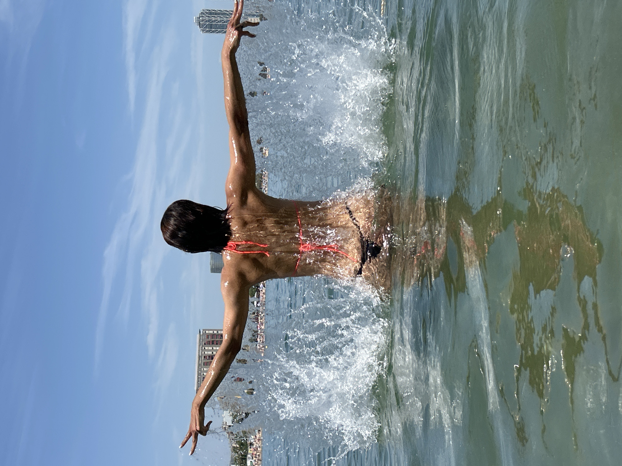 A person in a bright orange bikini with black bottoms is in the Mediterranean Sea, creating a large splash with their arms extended to the sides. Buildings and a crowded beach are visible in the background, indicating a location in the city of Barcelona, Spain, likely near the Barceloneta beach area due to the urban skyline and density of people. The sky is clear and blue with scattered white clouds. The water is green and reflective.