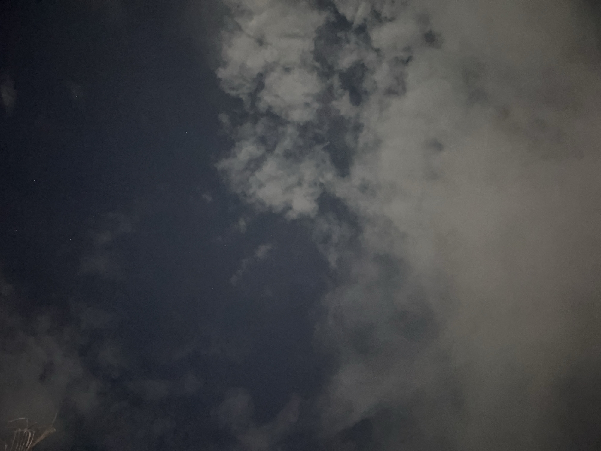 The image shows a nighttime view of the sky, likely in Canggu, Indonesia. A significant portion of the sky is obscured by clouds of varying density, with some areas appearing almost completely covered, while others reveal patches of dark, clear sky. A few faint stars are visible in the clearer sections. The overall lighting suggests it is either dusk or night with ambient light reflecting off the clouds. Part of what seems like a rooftop and structure is visible in the bottom left corner of the image.