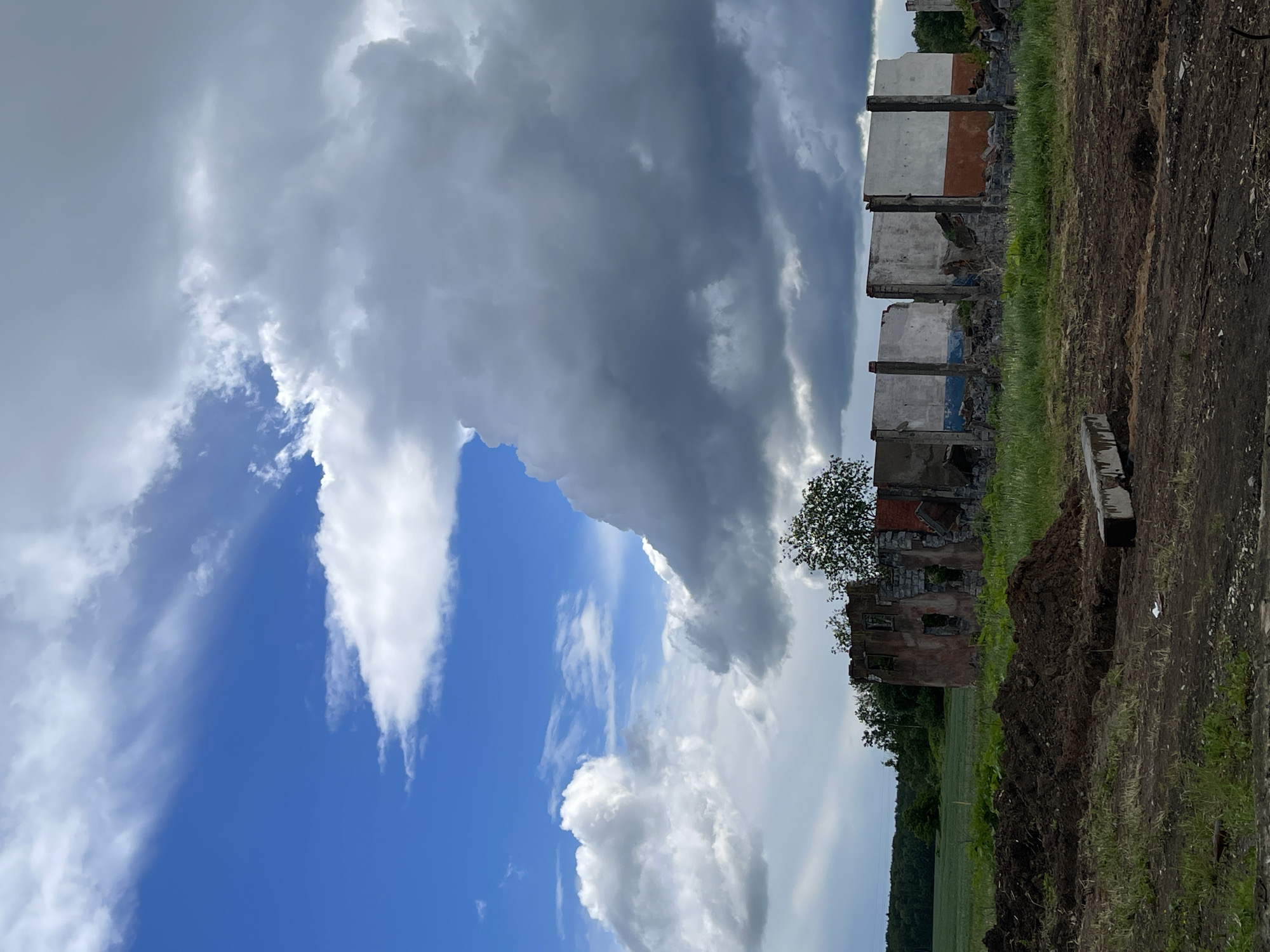 The image shows a partially ruined building with only remnants of walls remaining. The building appears to be in Киясово, Russia, as indicated by the location. Debris is visible around the structure. The foreground consists of dirt and grass. The sky is divided between blue and white clouds and large, looming gray storm clouds. A small tree is growing near one section of the dilapidated structure.