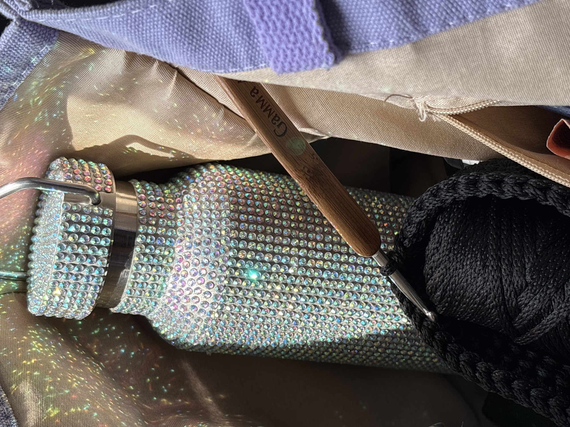 The image displays a close-up of a bedazzled water bottle and crochet supplies inside a bag. The water bottle is covered in numerous small, reflective rhinestones that create a shimmering effect, enhanced by light glinting off their surfaces and projecting colorful reflections onto the surrounding fabric. A crochet hook with a wooden handle, etched with the word "Gamma," is positioned in the foreground, with its metal hook engaged in black yarn. The yarn is wound into a thick, coiled shape. The bag appears to be made of different fabrics, including a beige material and a light purple fabric with a textured band. The scene is lit in a way that highlights the sparkle of the bottle and the textures of the materials. There is no discernible information in the image to indicate a specific location in Сухарэўскі сельскі Савет, Belarus.