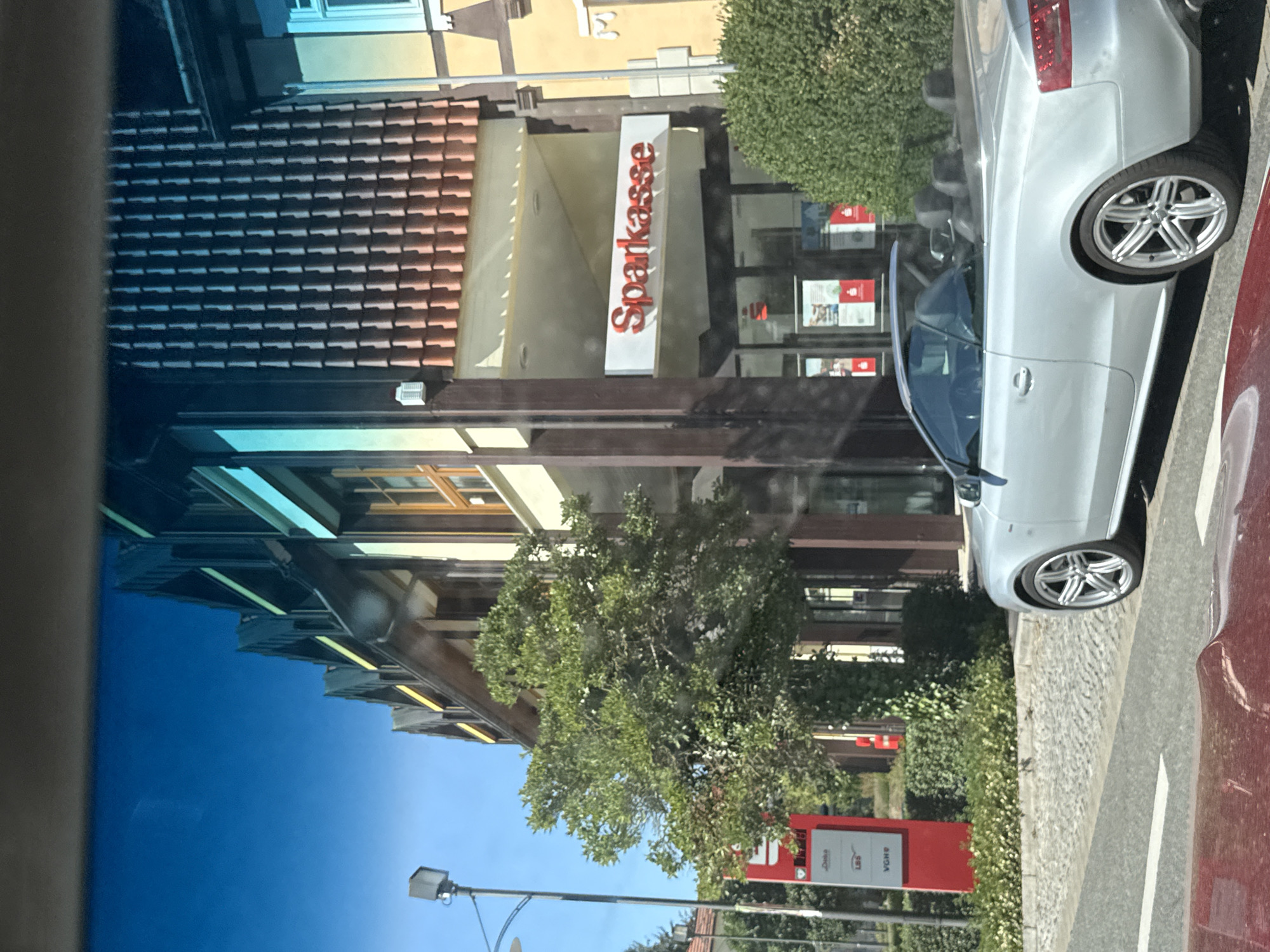 This is a photograph taken from inside a vehicle, looking out at a street scene in Bad Sachsa, Germany. A silver convertible is parked on the right side of the street, facing away from the camera. In the background, there is a building with a tiled roof and a sign that reads "Sparkasse". The building appears to be a bank or a financial institution. A tall red sign with white lettering is visible on the left side of the street, possibly indicating local businesses or services. A street lamp stands on the far left. The sky is clear and blue. The overall scene depicts a sunny day in a town.