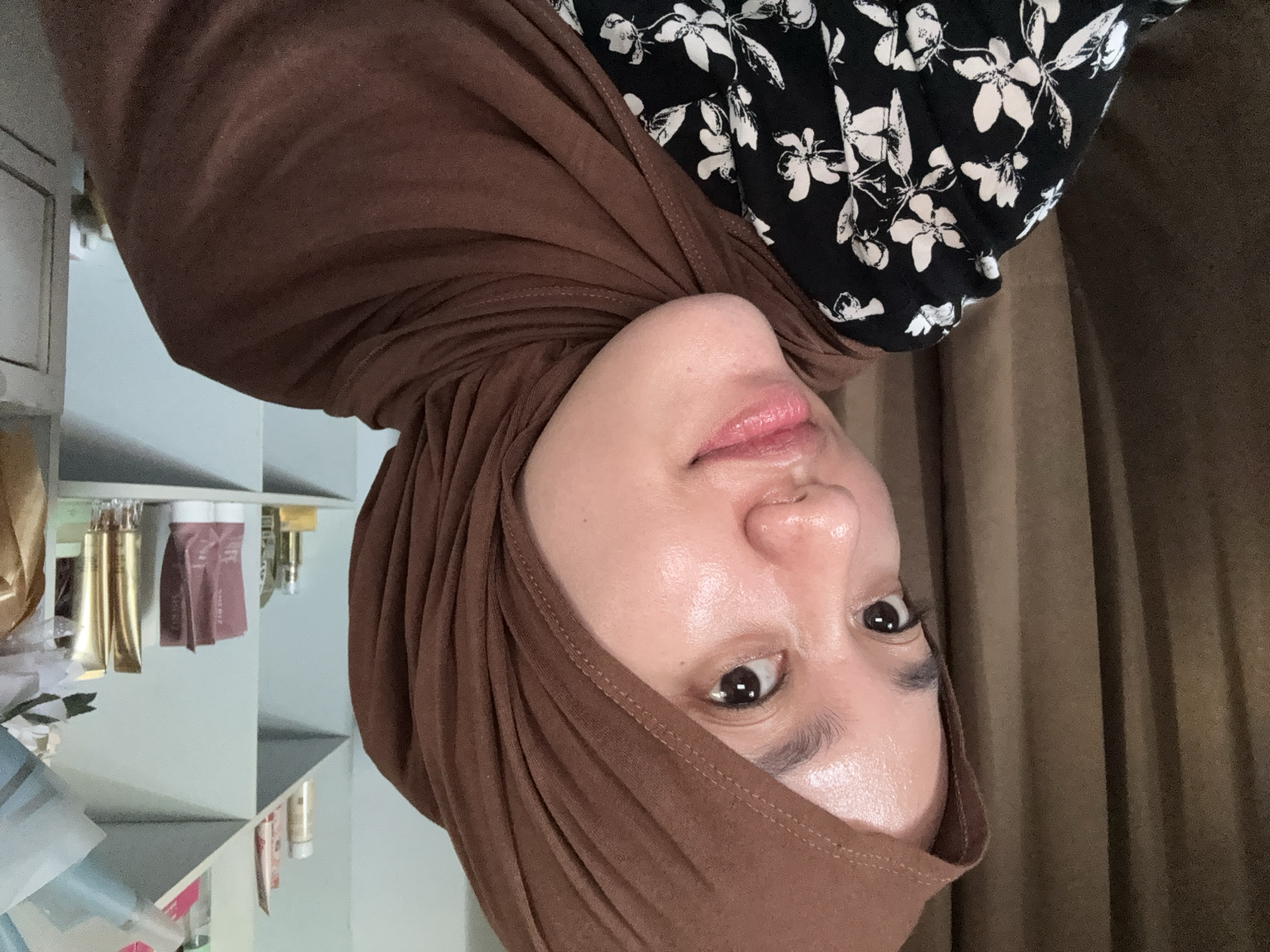 A person with a brown hijab is looking at the camera. The person is wearing a black and white floral shirt. There is a shelf with various cosmetic bottles. The background is a brown curtain. This is likely an indoor setting. No further details about the city can be determined from the image.