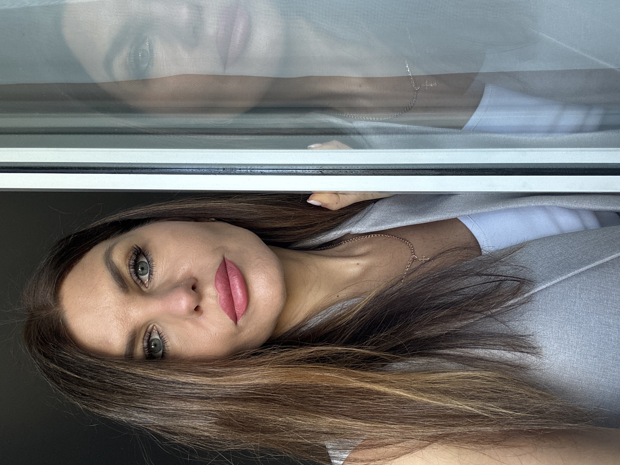 A woman with light skin, brown hair, and grey eyes is pictured indoors, partially visible with a reflection of her face above her. She is wearing a grey blazer over a white shirt and a delicate gold necklace. The upper portion of the image shows a window, reflecting the woman's face, neck, and a portion of her clothing. The scene is likely in a residential setting. There is no verifiable information to determine the specific location in Chaikovsky, Russia.