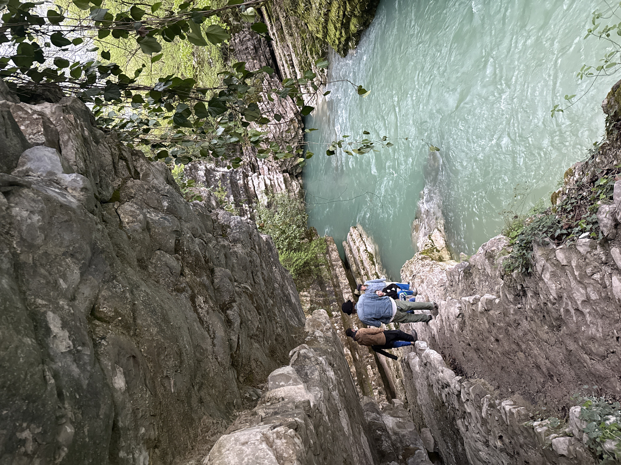 The image depicts a river gorge in Lebache, Georgia. The gorge walls are composed of jagged, layered rock formations, dropping sharply to a turquoise-colored river below. Two individuals are positioned on the edge of the gorge. One is wearing a denim jacket, dark pants, and blue shoes and is lying with their head towards the water. A second person is standing beside them in a brown jacket, black pants, and blue shoes. The viewpoint is looking down into the gorge, capturing both the rock structure and the water. Some foliage and trees are visible along the top edge of the gorge.