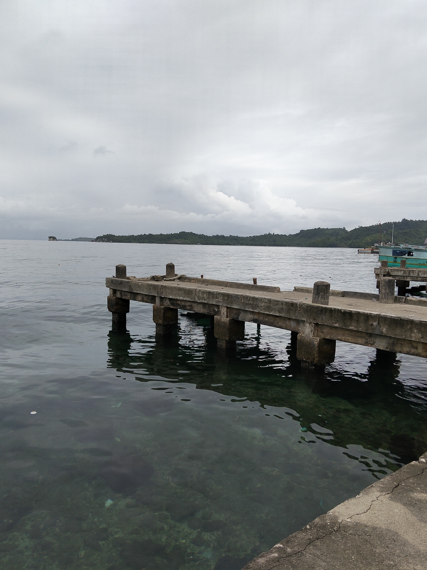 A weathered concrete pier extends into calm, clear water under an overcast sky. The pier is supported by thick concrete pillars and has a rough, uneven surface. In the distance, a chain of lush green islands with dense vegetation lines the horizon. On the right side of the image, a portion of a green and blue boat is docked at a separate pier. The water is transparent enough to reveal a rocky seabed beneath. The scene appears to be located in Banggai, Indonesia, given the distinct tropical island landscape. There are no people visible in the frame.