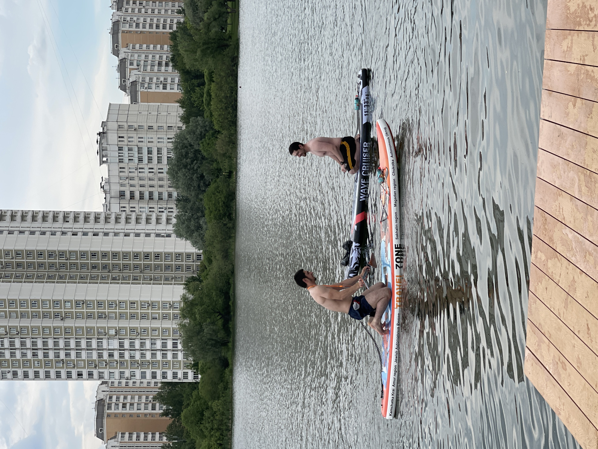 The image captures a daytime scene of two shirtless men paddleboarding on a wide body of water, likely a reservoir or lake, in Moscow, Russia. Both men are kneeling on their respective stand-up paddleboards (SUPs), suggesting they are either initiating their activity, paddling on their knees, or taking a moment to rest.
The man on the left is wearing dark shorts and is positioned towards the front of his paddleboard, holding a paddle. His board is predominantly orange, white, and blue, with visible text including "TRAVEL ZONE", "Kamchatka Krai", "Amur region", "Jewish Autonomous Region", and "Sakhalin region". The man on the right, wearing black and yellow shorts, is also kneeling, and a portion of his paddle is visible. His paddleboard is black and white, featuring the text "WAVE CRUISER", "11' 33"6"", "Magadan region", and "Chukotka Autonomous Okrug".
In the background, a dense line of green trees separates the water from a prominent urban landscape. Numerous tall, multi-story residential buildings, consistent with Moscow's architectural style, dominate the distant shoreline. A wooden boardwalk or dock is partially visible on the right edge of the foreground, suggesting a launching or resting point for water activities. The sky is overcast, indicating a soft, diffused natural light, typical of a cloudy day. The overall atmosphere appears tranquil and recreational. The names of various Russian regions visible on the paddleboards serve as a strong geographical cue, reinforcing the provided location context of Moscow, Russia.