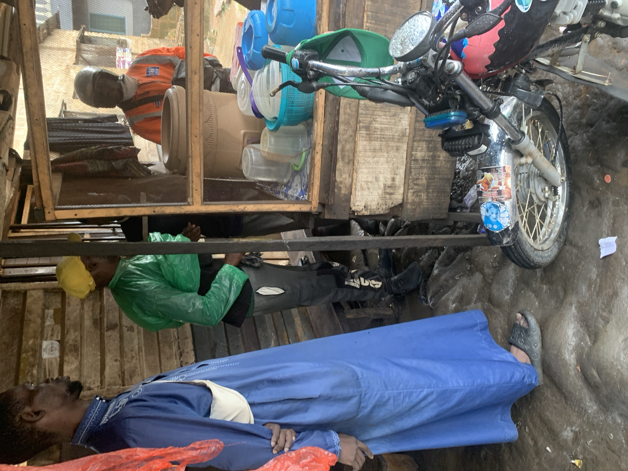 The image depicts a scene outdoors, likely in Shagamu, Nigeria, characterized by a rustic wooden structure that appears to be a stall or shop. The ground is muddy and uneven. In the foreground, a motorcycle is parked, its front wheel and handlebars visible. The motorcycle is decorated with stickers.
Three individuals are present. On the left, a person wearing a long, flowing blue garment with a white pouch across their chest stands facing away from the camera, looking towards the right. Next to them, a person wearing a bright green, hooded jacket and dark pants is also facing right, partially obscured. Further back and to the right, inside the wooden structure, a person wearing an orange high-visibility jacket and a helmet with a visor is visible, appearing to be engaged in some activity within the stall. The stall itself is open to the front, with shelves displaying various plastic containers, buckets, and other items. The overall environment suggests a market or roadside vending area. The lighting indicates it is daytime.