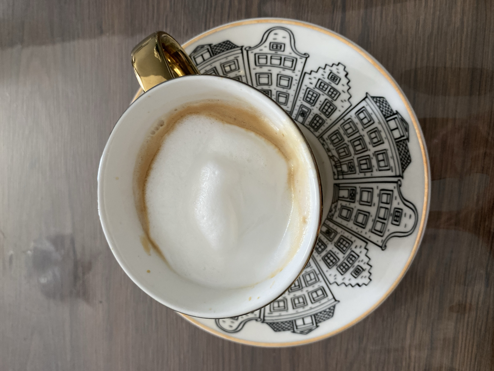 An overhead photograph showcases a white coffee cup filled with a frothy, milky coffee beverage, likely a latte or cappuccino. The cup has a distinct gold-colored handle and a thin gold rim. It is placed on a matching white saucer, which is intricately decorated with black line drawings of several stylized buildings. These buildings feature gabled roofs and numerous windows, characteristic of traditional Dutch canal houses or similar European architecture.
The coffee drink in the cup has a thick layer of creamy white foam, with visible streaks of light brown coffee along the inner rim of the cup. The cup and saucer are resting on a warm, brown wooden surface, possibly a table, which exhibits a subtle wood grain pattern and some light reflections. The lighting is soft and even, highlighting the details of the ceramics and the coffee.
The scene suggests a quiet moment of enjoying a beverage. While the architecture on the saucer hints at a Dutch design aesthetic, the image is located in Saint Petersburg, Russia, suggesting this could be a souvenir or a decorative item found in the city. There is no visible text on the cup, saucer, or surrounding surface. The image conveys a sense of calm and simplicity.