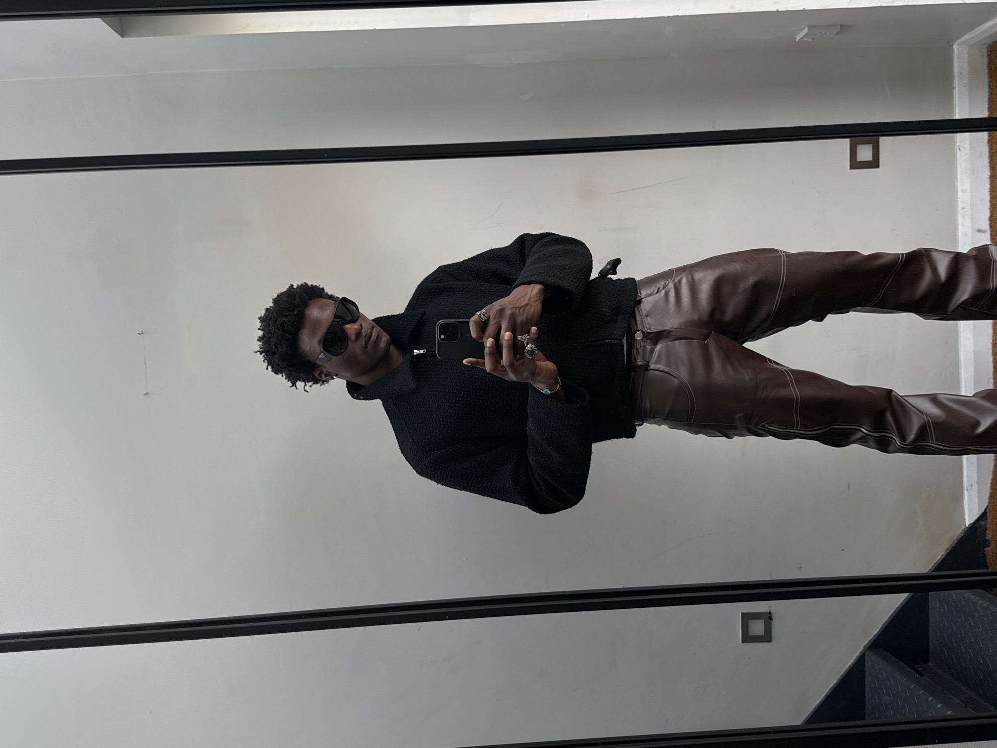 A slender, dark-skinned individual with short, curly black hair is centrally positioned, facing slightly left, in front of a mirror. The subject is wearing sunglasses, a dark, textured jacket, and brown leather trousers with visible seams. The individual is holding a black smartphone and seems to be taking a selfie. The background consists of a plain white wall with several square-shaped objects mounted on it. The scene includes a portion of a staircase with metal treads, visible in the bottom right corner. The location is likely an interior space within a building. Specific location in London not evident from the image.