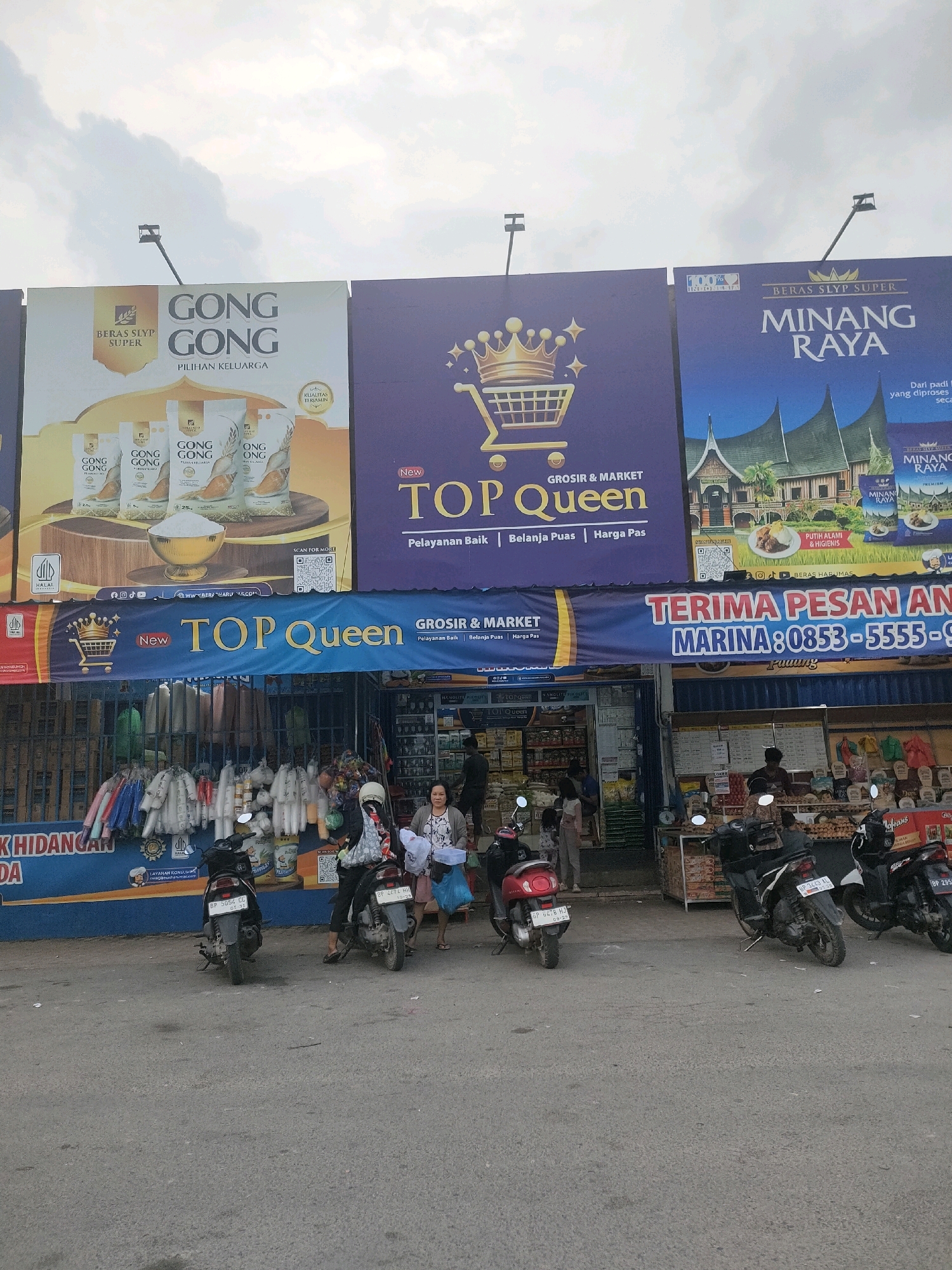 The image depicts a street scene in front of a market or shop in Batam, Indonesia. Three large billboards are prominently displayed above the storefront. The leftmost billboard advertises "GONG GONG BERAS SLYP SUPER" with images of rice packaging. The central billboard features a large shopping cart icon and the text "TOP QUEEN GROSIR & MARKET". The rightmost billboard displays "BERAS SLYP SUPER MINANG RAYA" with an image of traditional Minangkabau houses.
Below the billboards, a banner reads "TERIMA PESAN AN MARINA: 0853-5555-9319". The storefront itself appears to be a grocery or convenience store with shelves of products visible through glass doors and openings. Clothing items are displayed hanging outside the storefront on the left. Several motorcycles are parked on the asphalt in front of the store, with a few people visible around the motorcycles and inside the store. The sky above is overcast. The overall scene suggests a commercial area with a focus on food products and local commerce.