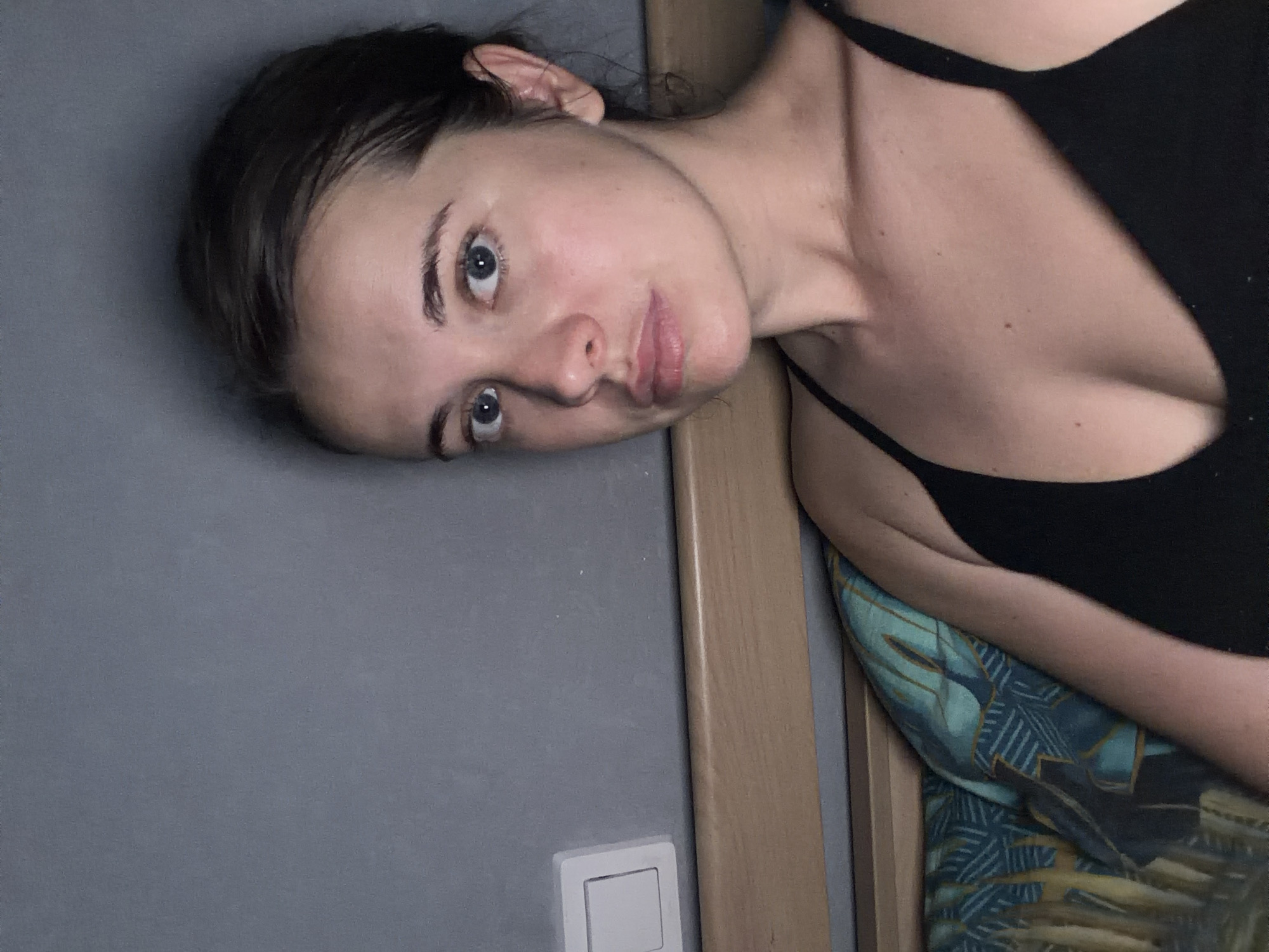 A young woman with dark hair, pulled back, and striking blue-gray eyes, looks directly at the camera in this indoor portrait. Her head is tilted, conveying a relaxed and direct gaze, with her lips slightly parted. She has fair skin and prominent eyebrows. She is wearing a black spaghetti strap top or bra, parts of which are visible, revealing her shoulder and upper chest.
The woman appears to be leaning against or lying beside a light grey wall. To her right, a wooden door frame runs vertically, suggesting a doorway or partition. On the grey wall, near the bottom left, a white rectangular light switch is visible. Below and to the right of the woman, a vibrant patterned fabric, possibly a pillowcase or bedding, featuring shades of blue, green, and gold with geometric designs, adds a splash of color to the scene. The lighting is soft and ambient, consistent with an indoor setting, and does not suggest strong natural daylight. The overall atmosphere is calm and intimate. The location context for this image is Sochi, Russia. There is no visible text in the image.